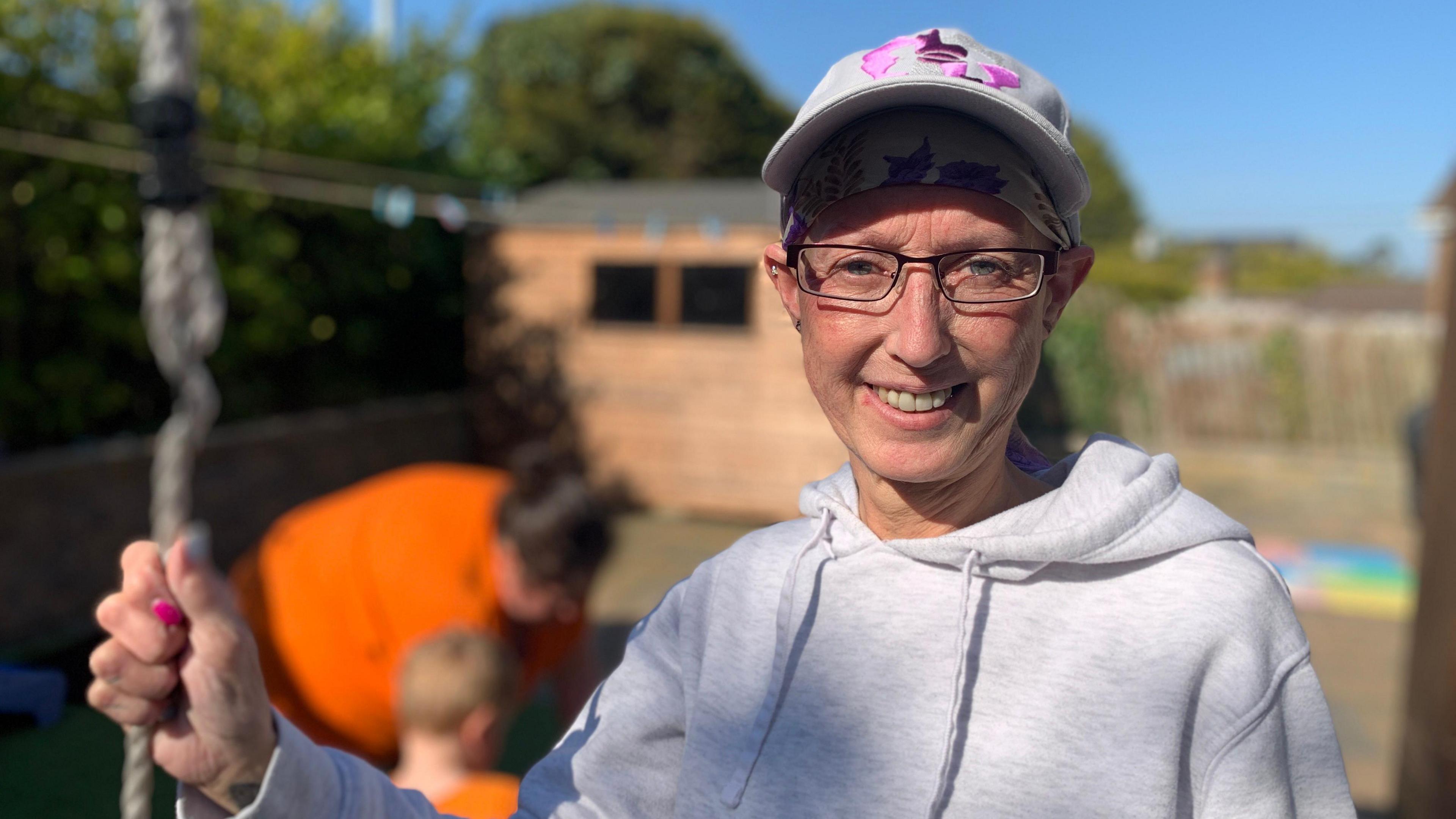 A woman in a grey hoodie smiles at the camera. She wears glasses, a baseball hate and holds a piece of playground equipment in her hand. IN the background you can see a child being helped by another woman but the background is blurred.