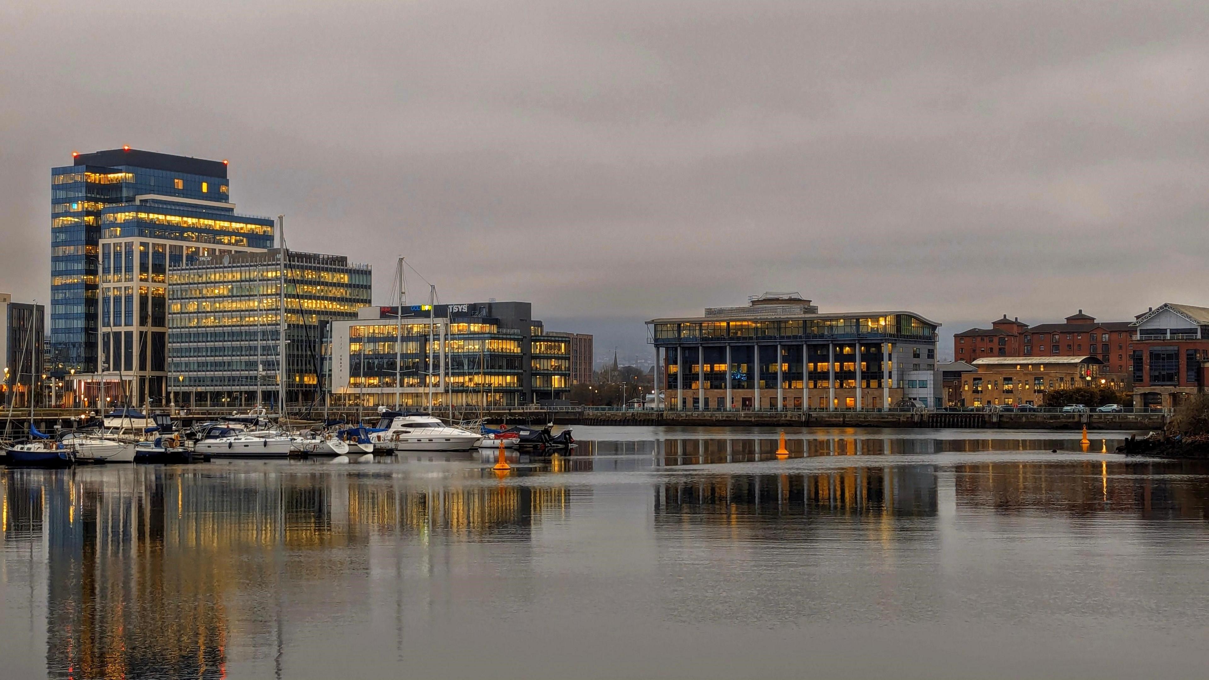 Belfast view from the Maritime Mile showcasing boats docked in the harbour and buildings lit up in the early evening. The sky in the background has a pink tone to it but clouds fill the sky. 