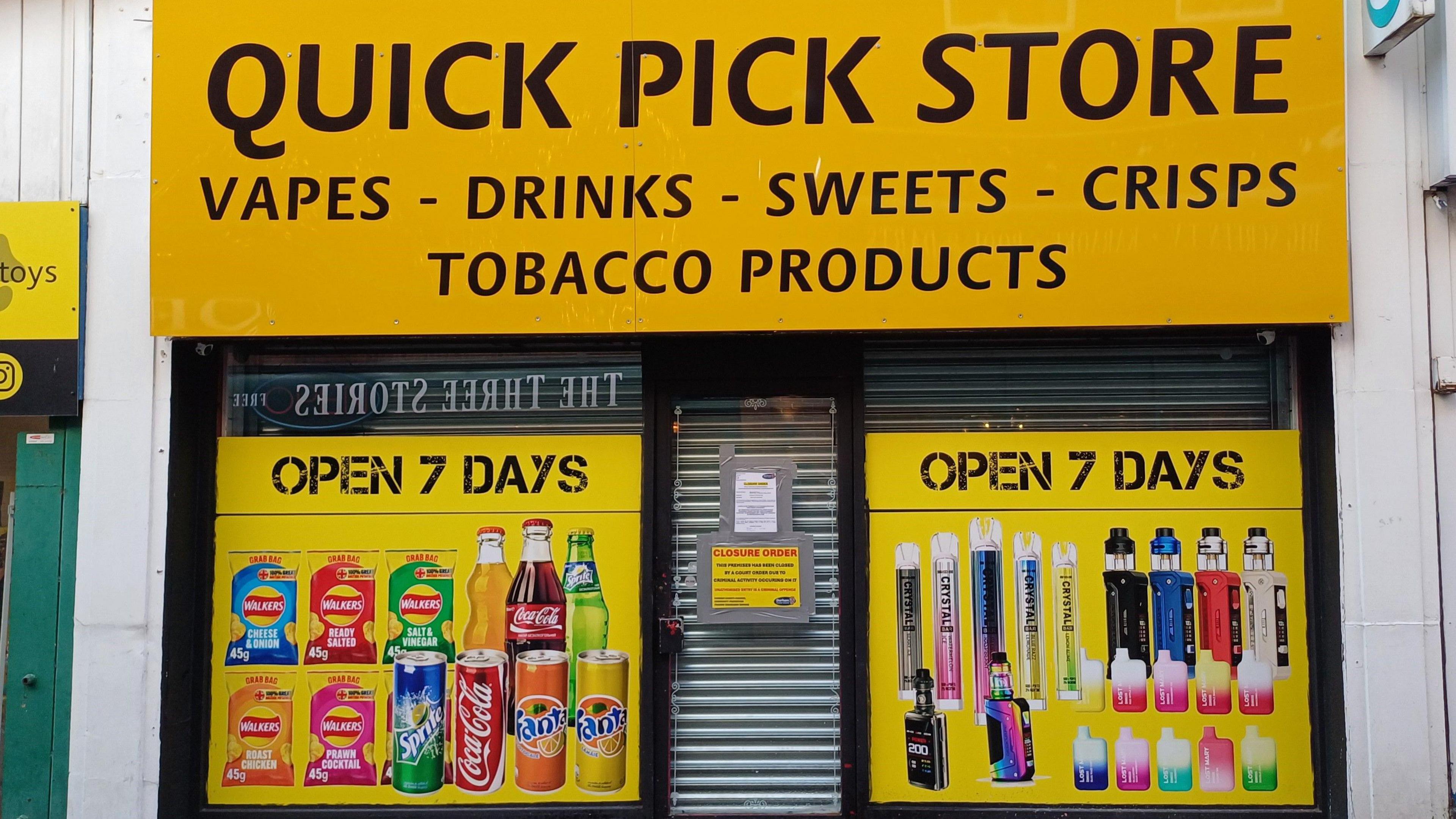 The outside of the Quick Pick Store which has a bright yellow front. The words 'vapes, drinks, sweets, crisps and tobacco products' is written above the door.