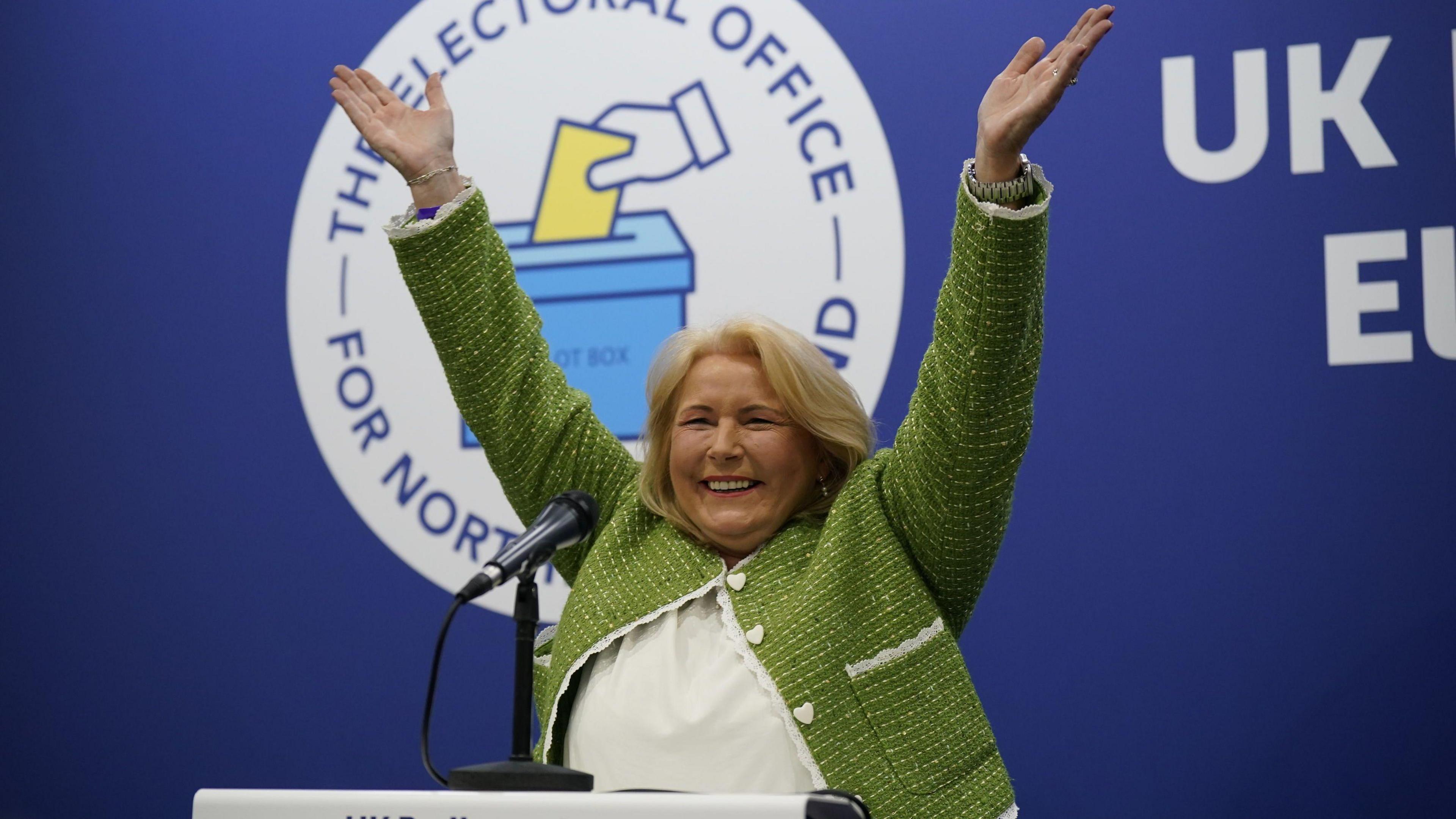 Pat Cullen throws her hands in the air and smiles as she becomes elected to Westminster in July 2024's UK general election. She has shoulder-length blonde hair, and is wearing a green coat with white stitching and heart-shaped buttons. She is also wearing a white shirt underneath. 