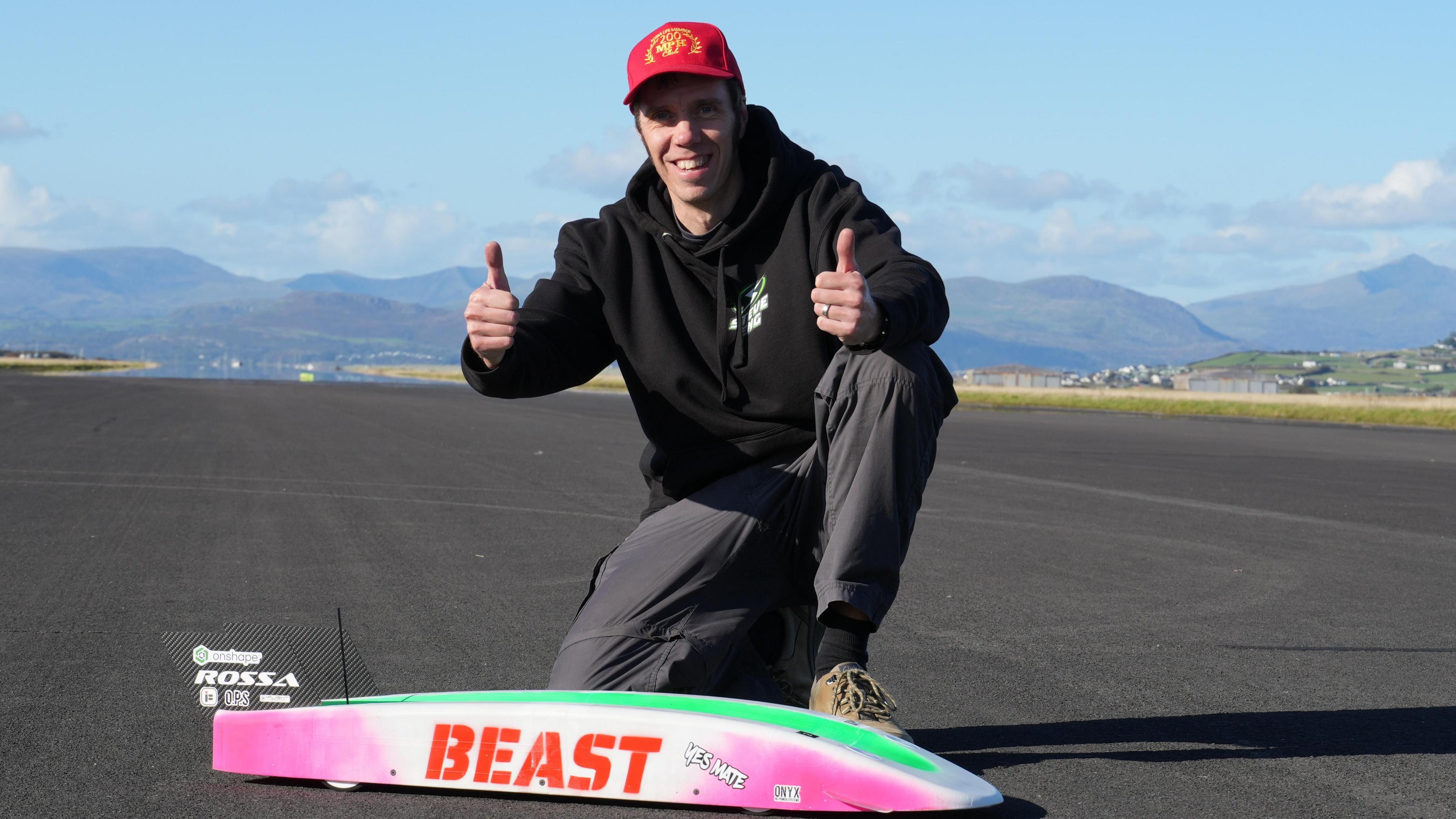 The image shows a man with a red baseball cap and a black hoodie, giving a double thumbs-up, kneeling behind a remote control car on a long stretch of road.