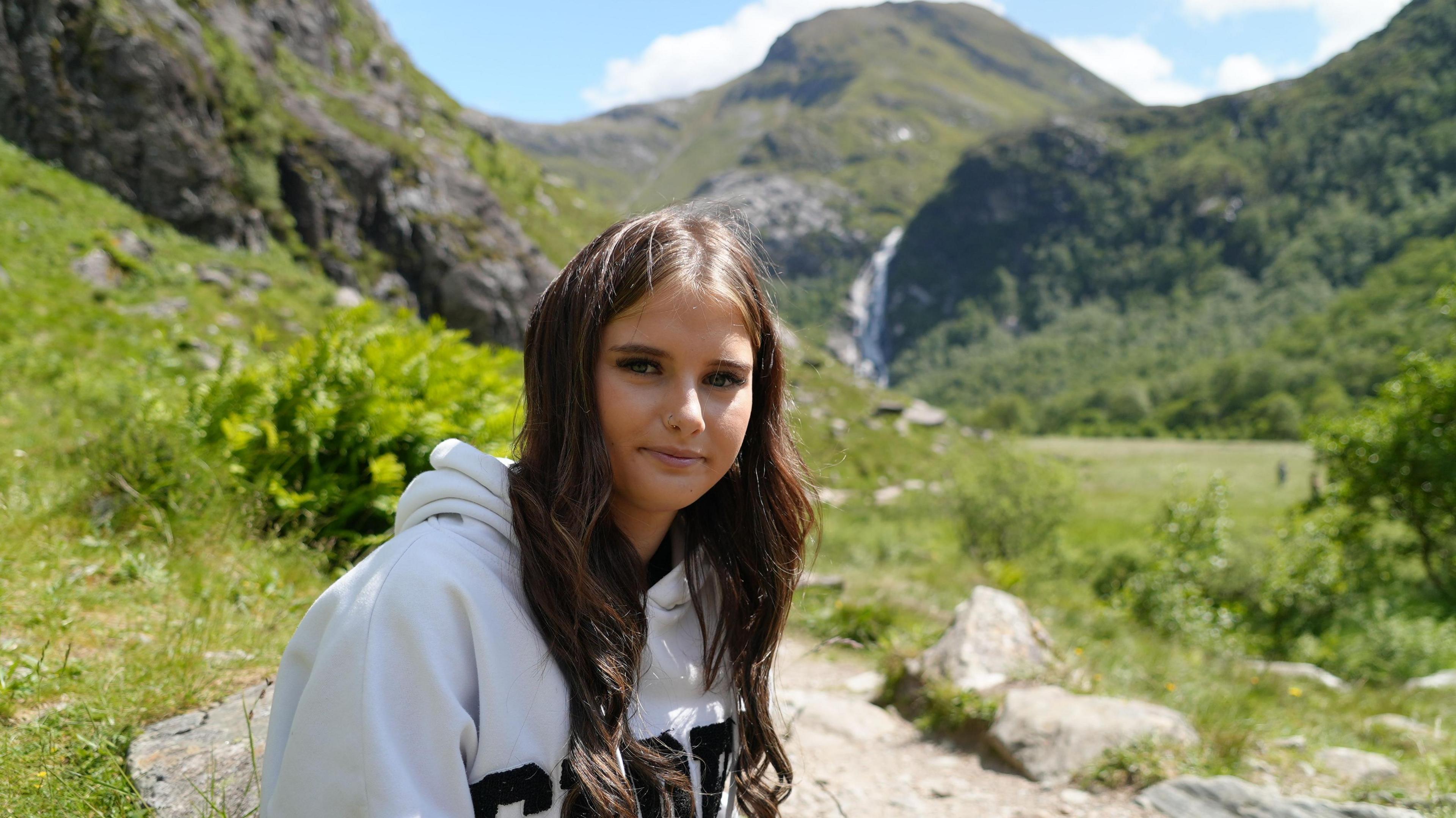 Ellie, who has long, brown hair and is wearing a grey hoodie, is sitting in a glen surrounded by fields and mountains