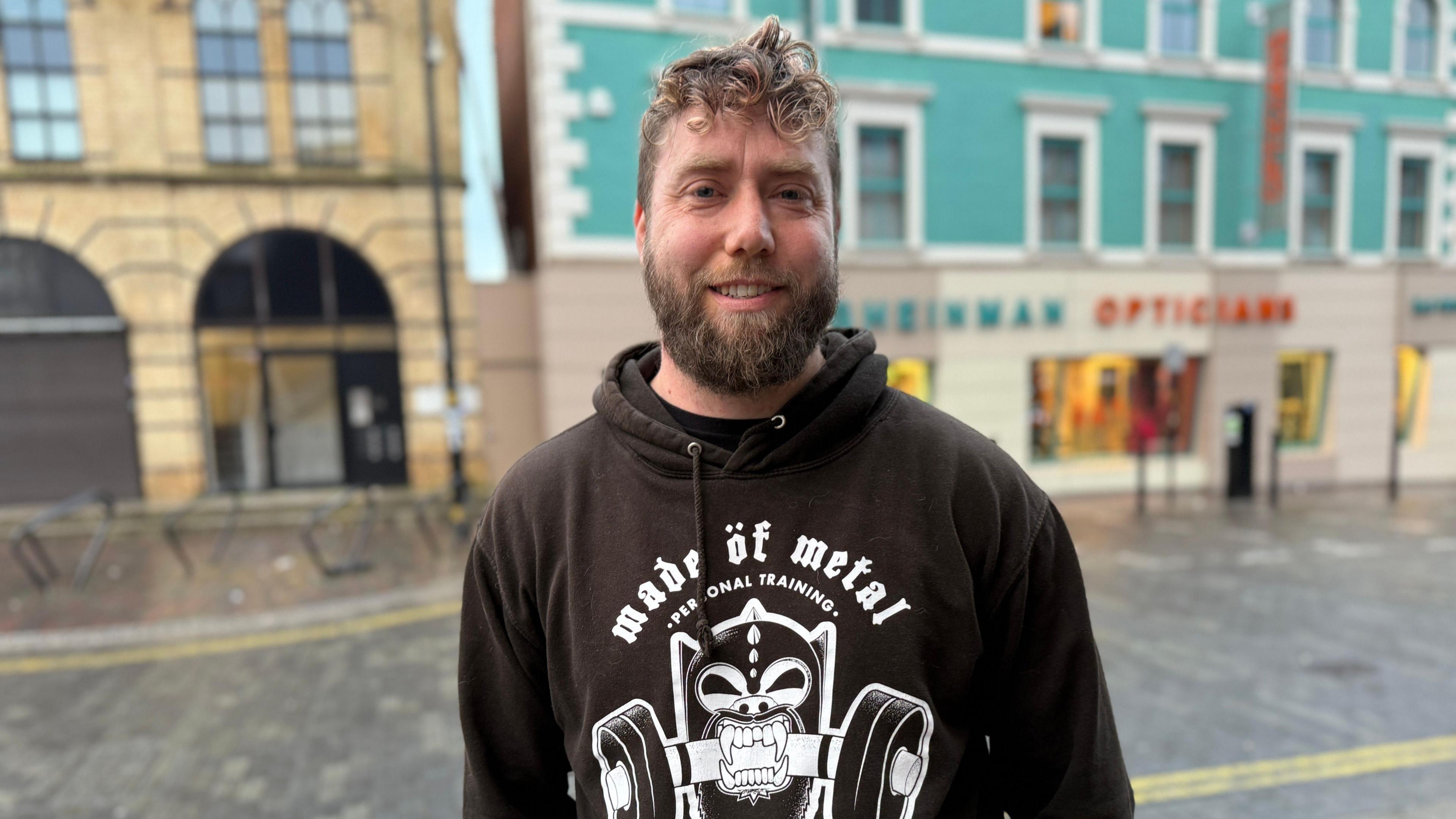 A man with curly light brown hair and a full beard stands in the town centre. He is wearing a black hoodie with the Made of Metal logo on it. 