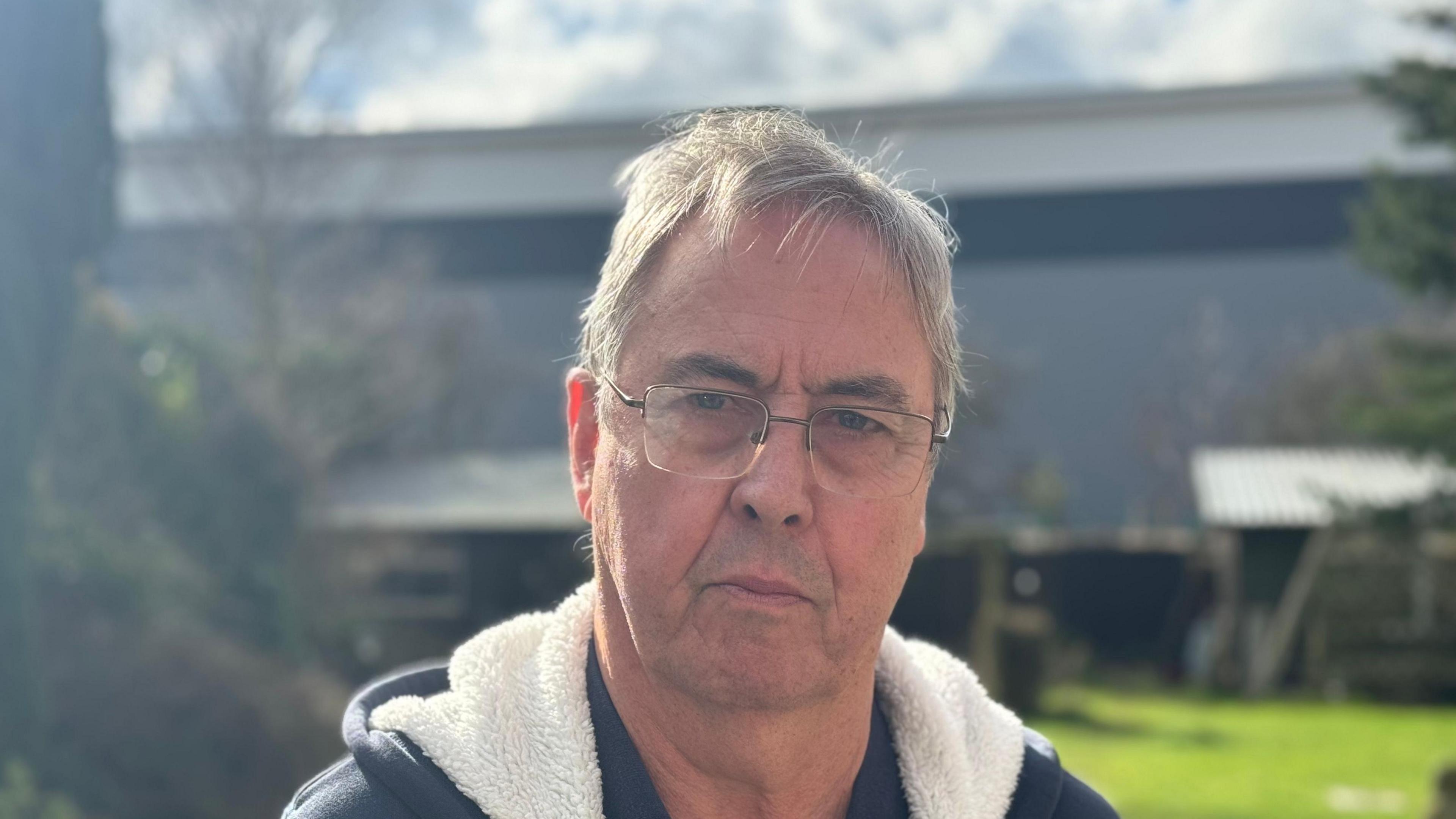 Dave Ward -  a man wearing glasses and a navy and cream hoody standing in a garden staring into the camera. The warehouse can be seen in the background.