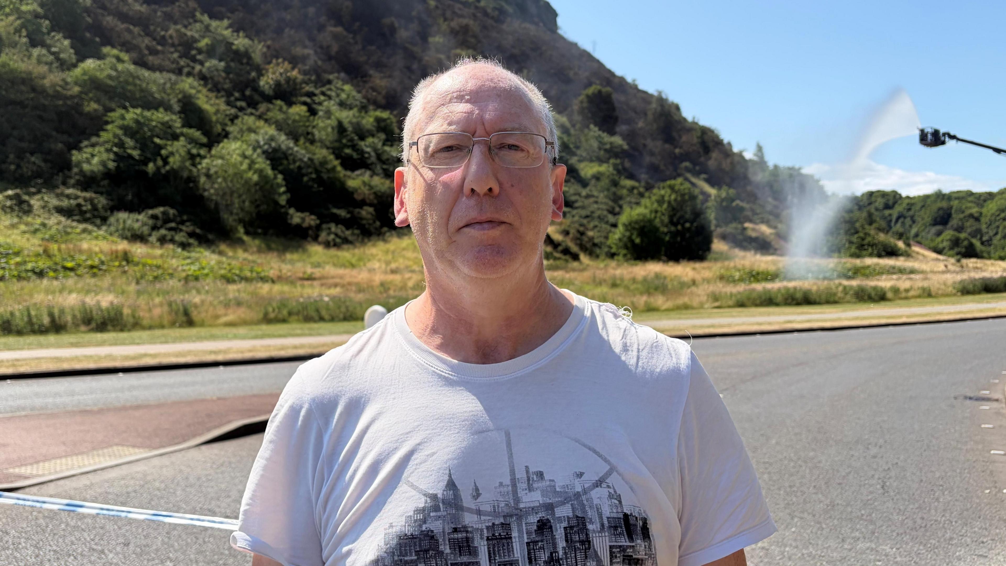 Callum Smith standing in from of Arthur's Seat. He is wearing a white t-shirt and glasses. Firefighters are hosing the hill down in the background.