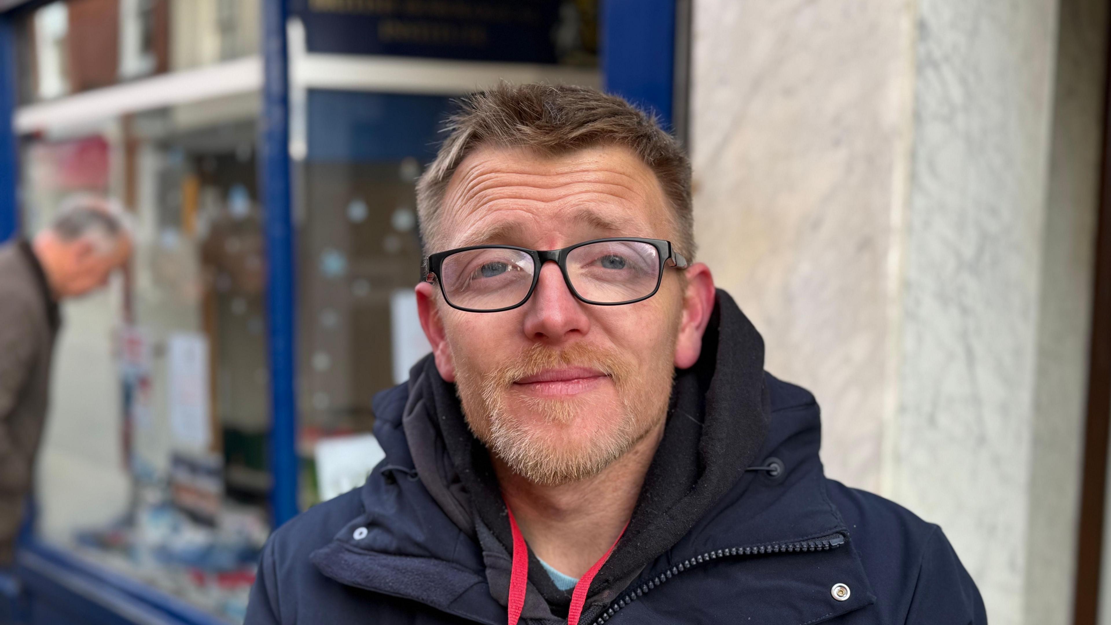 A man with short light brown hair and facial hair and glasses staring into the camera. He is wearing a navy blue jacket with a hood.