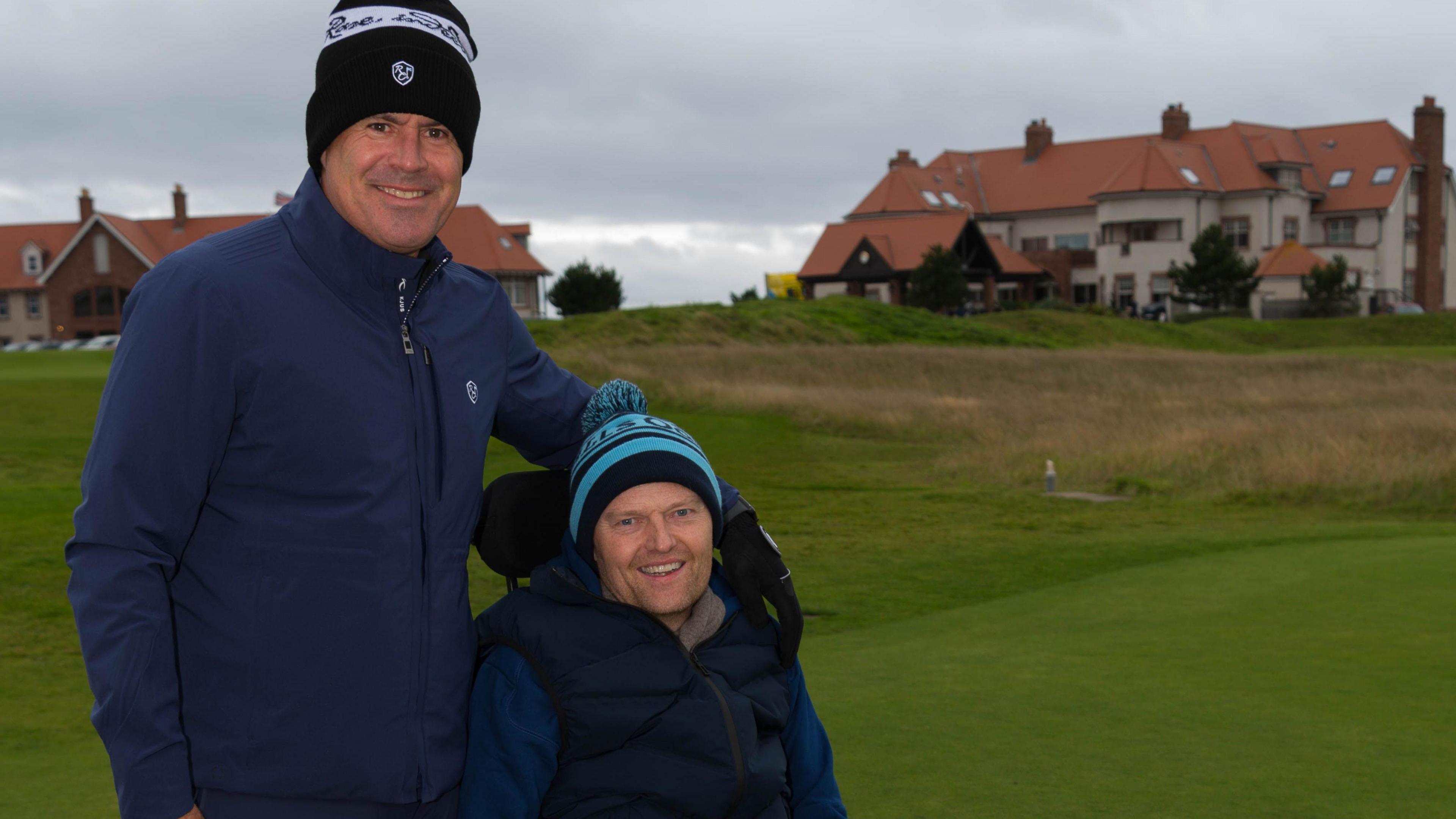 Kenny is standing up with his arm around Scott's shoulders. scott is sitting down in a wheelchair. They are both wearing wooly hats and are on a golf course.