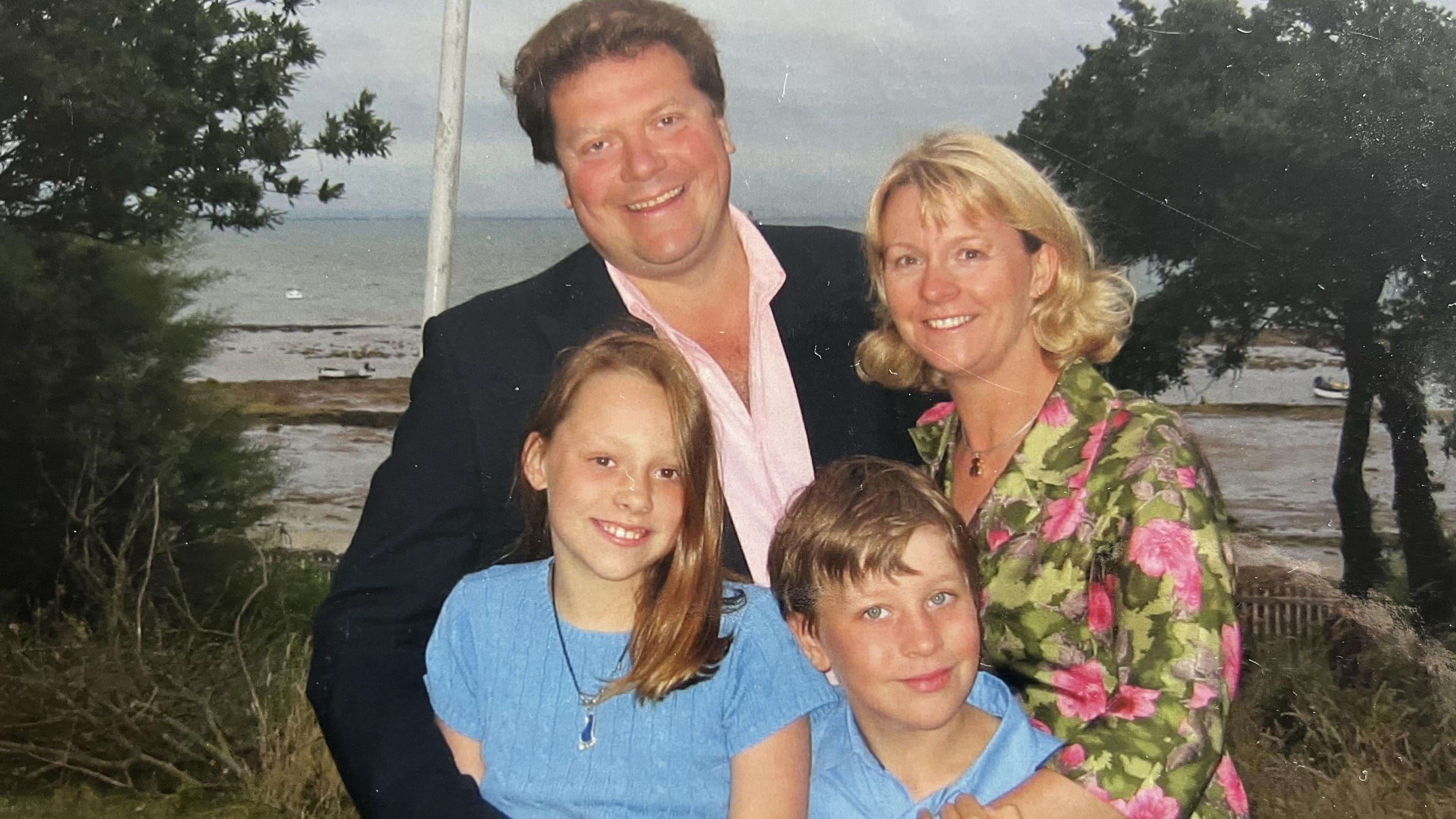 An older Alex and Sue stand with two children in front of them, they all smile at the camera in front of water, trees and grass. Alex wears a black suit jacket and pink shirt, Sue wears a pink flowered dress with green leaves. The children wear blue.