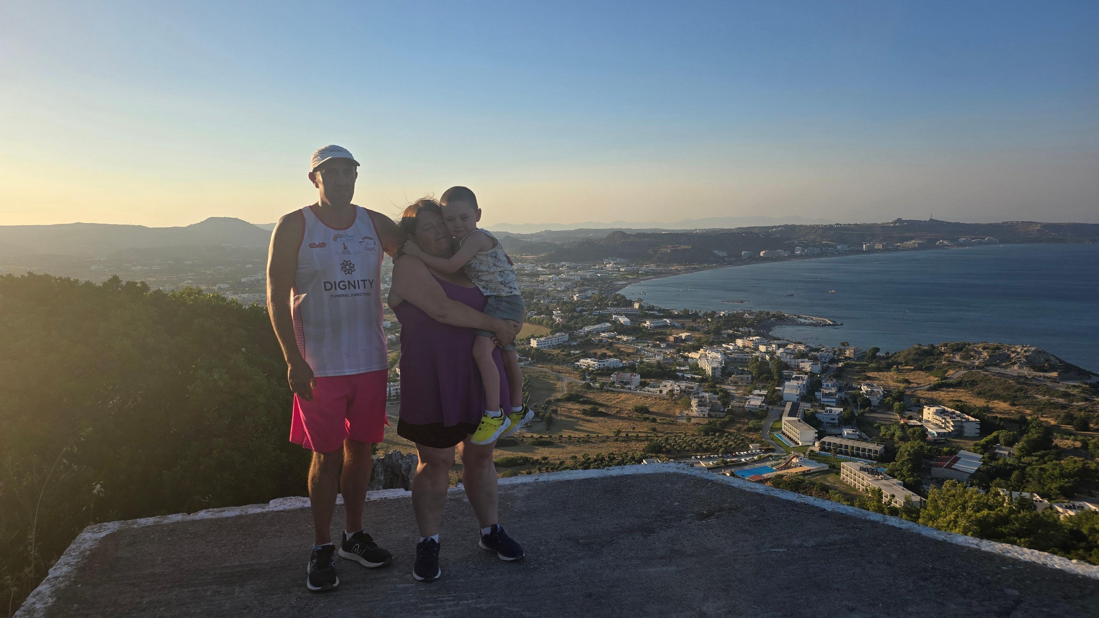A family of three stand atop a platform against the backdrop of a coastal setting at sunset. The man is wearing a white baseball cap, vest and pink shorts. The woman is wearing a purple top and black shorts and is holding a small boy of about five years old. The sea and a smattering of white properties stand behind them.