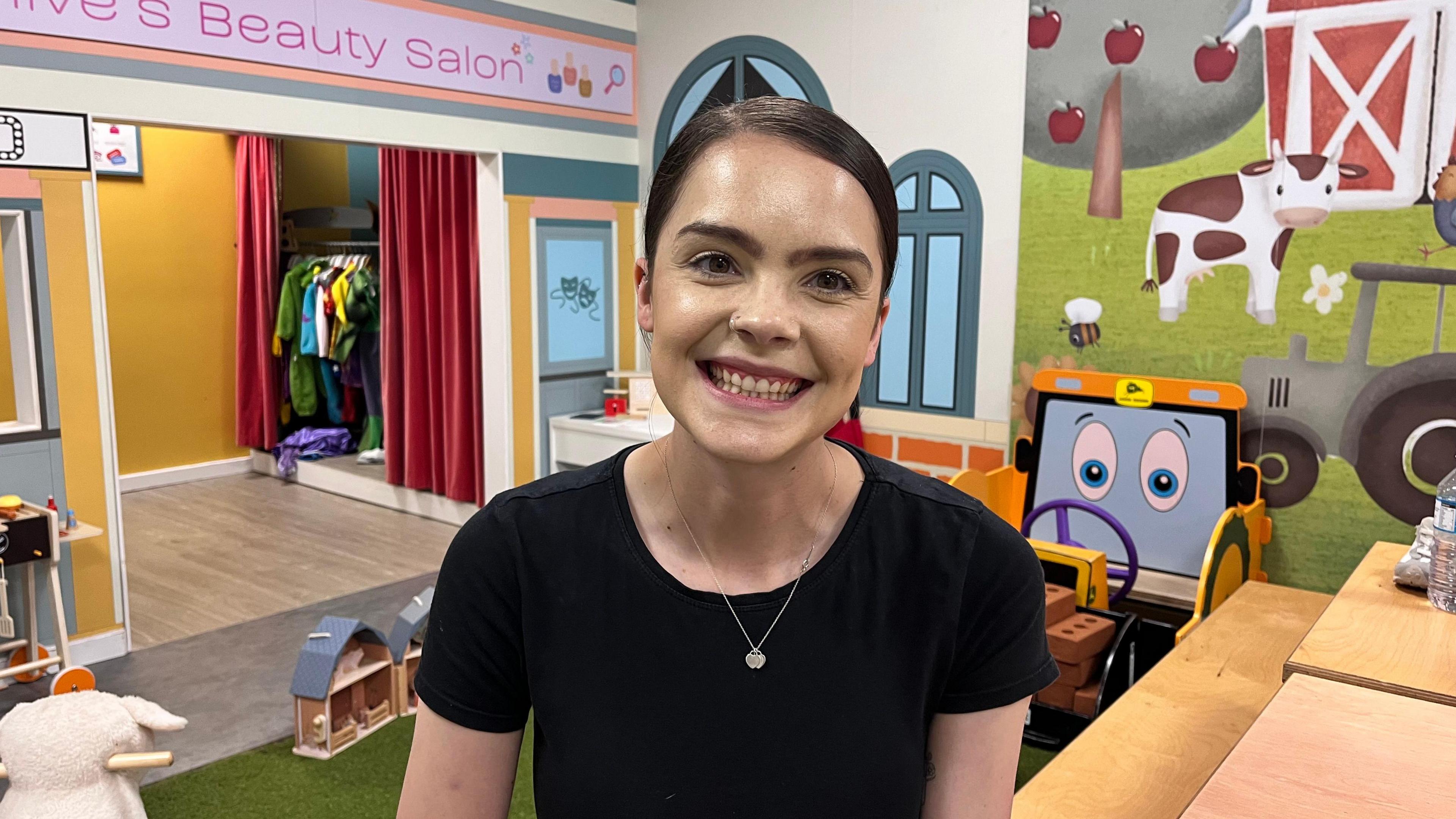 Lily Kemp sits in an indoor child's play centre in Hull smiling to the camera wearing a black t-shirt with toys and children's dressing up outfits in the background.