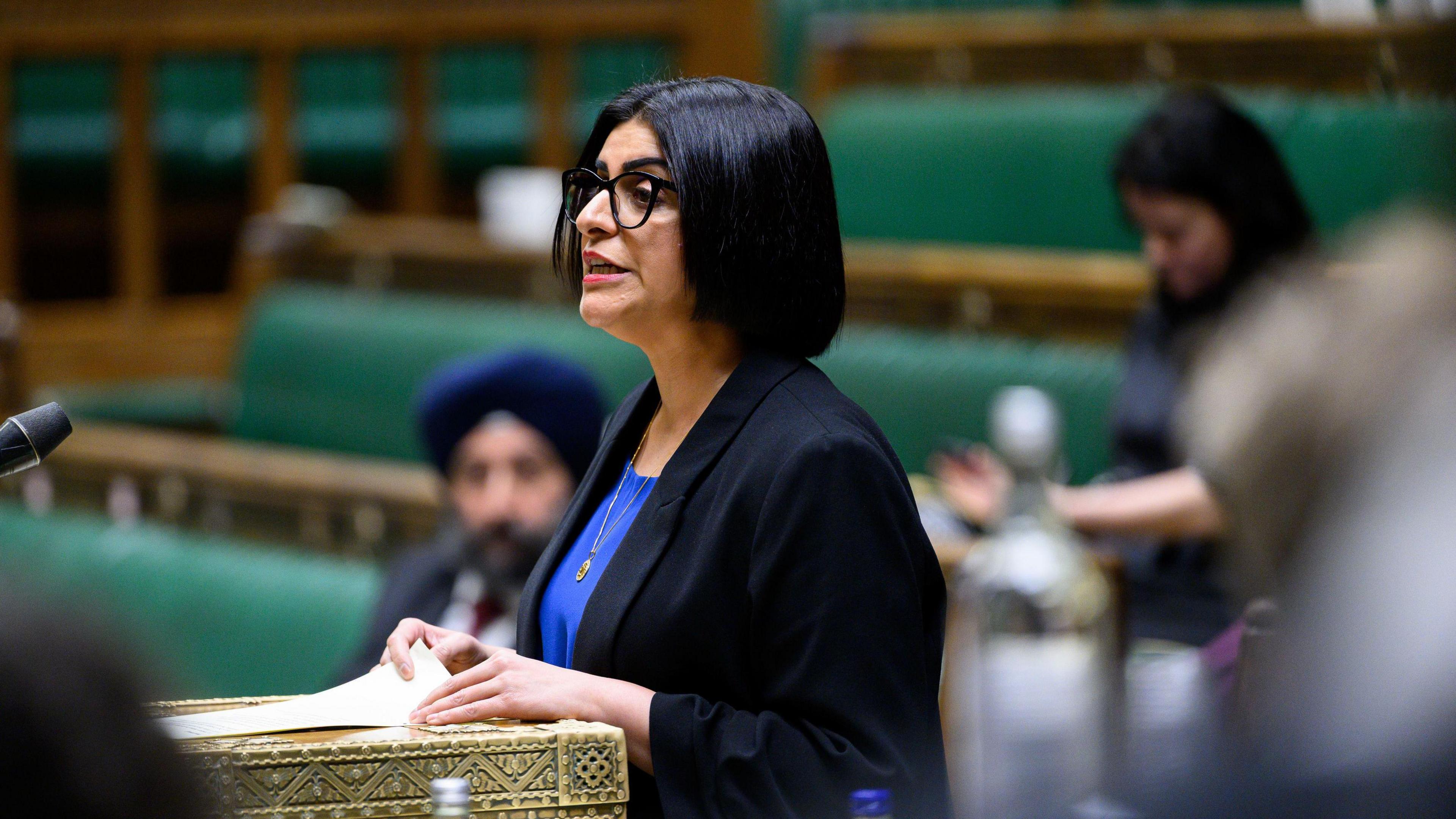 Home Secretary Shabana Mahmood in the Commons. She has shoulder-length black hair and is wearing a black suit jacket and blue shirt. A few other MPs are visible in the blurred background.