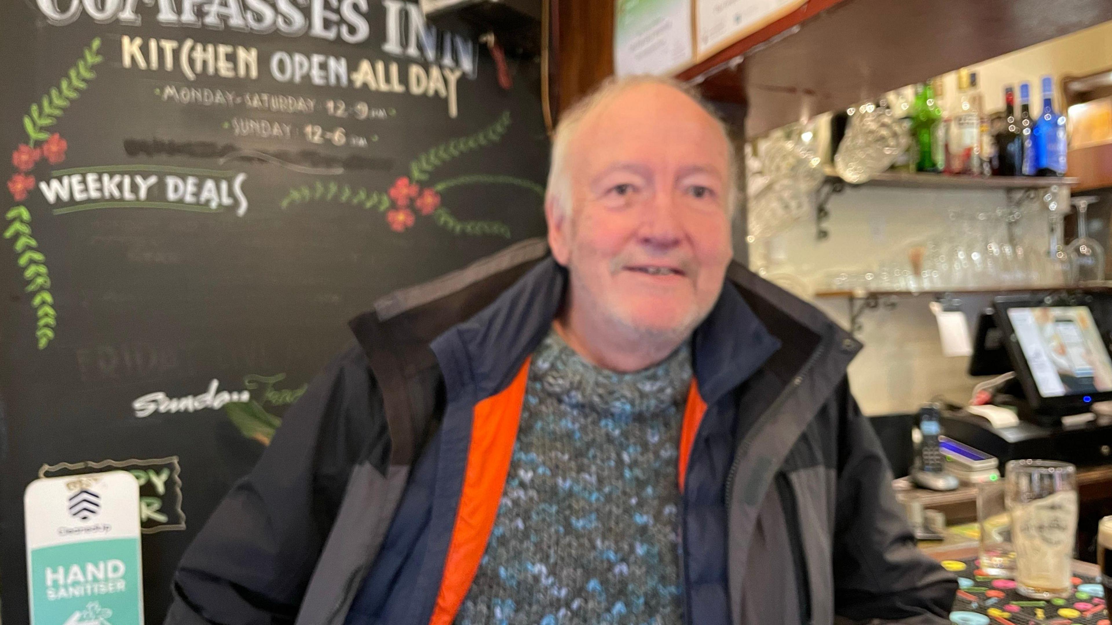 A man with a woolly jumper and two coats on stands at a bar in a pub