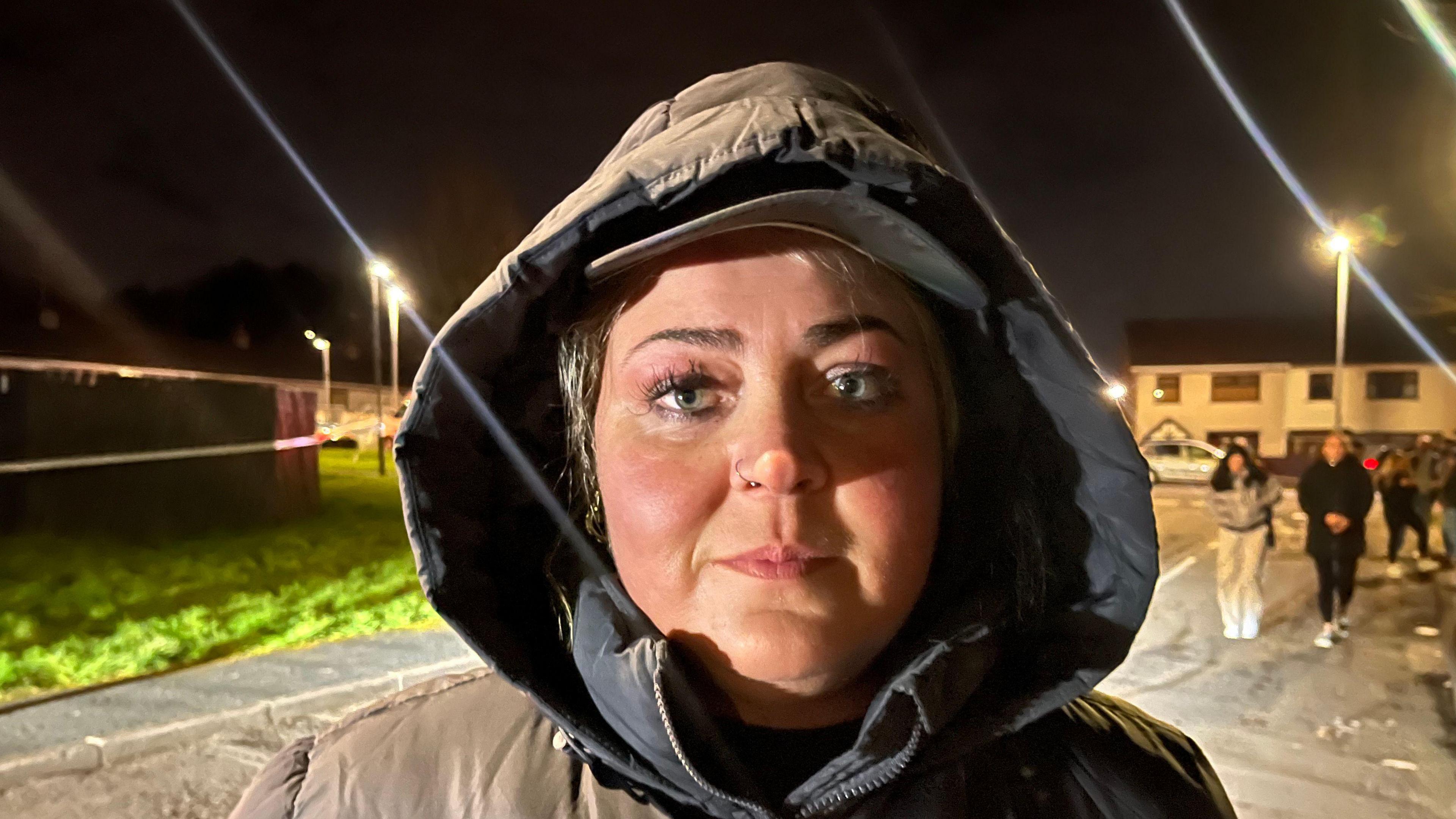 A woman wearing a green/grey coat with the hood up. She is staring into the camera and has fair hair.