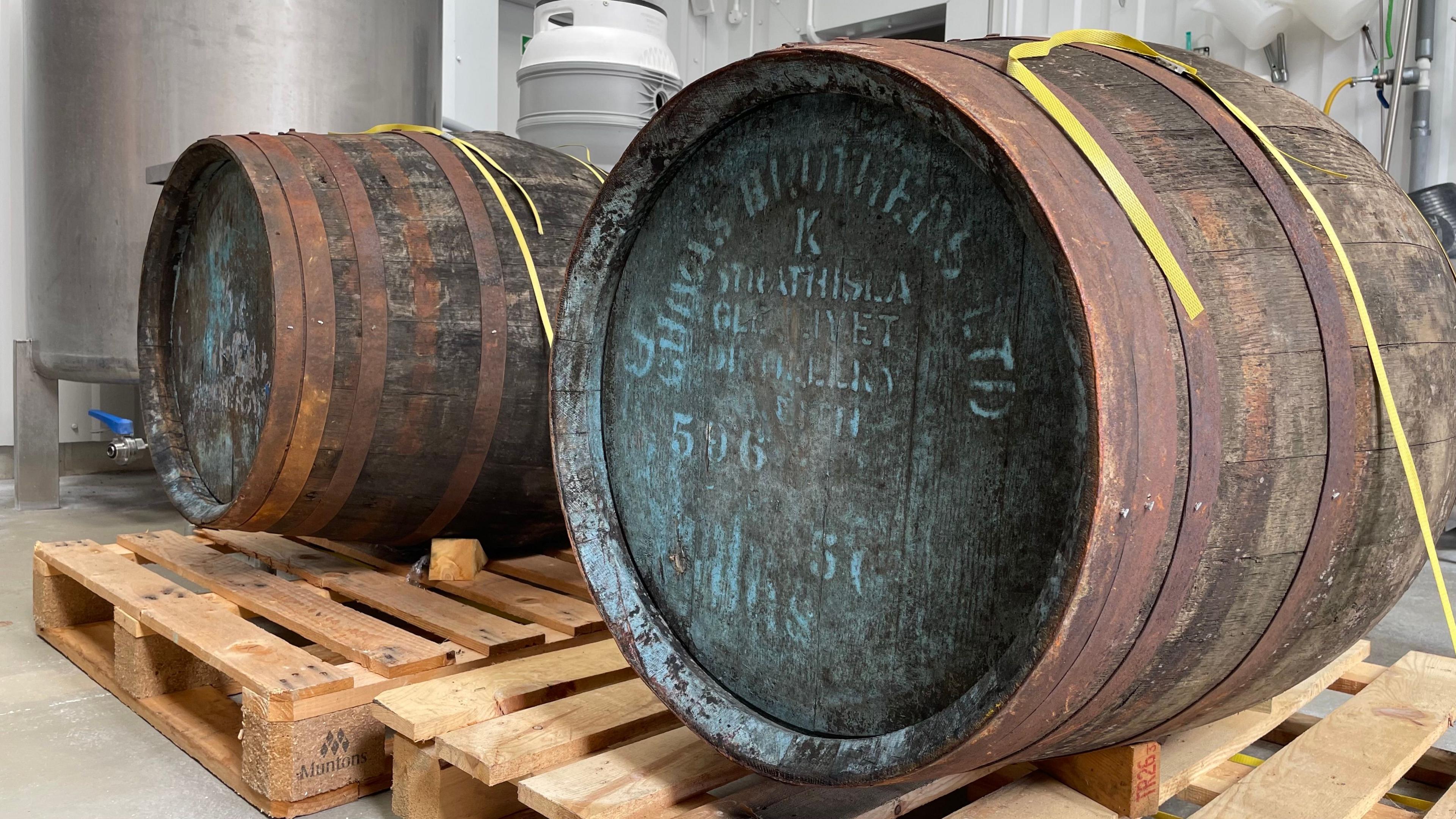 Two large, old wooden barrels sitting on top of two pallets. They are both inside the brewery's brewhouse. They are dark in colour and have blue writing on their lids, though they are both lying on their sides. Yellow strapping is holding them in place. Brewing equipment can be seen in the background.