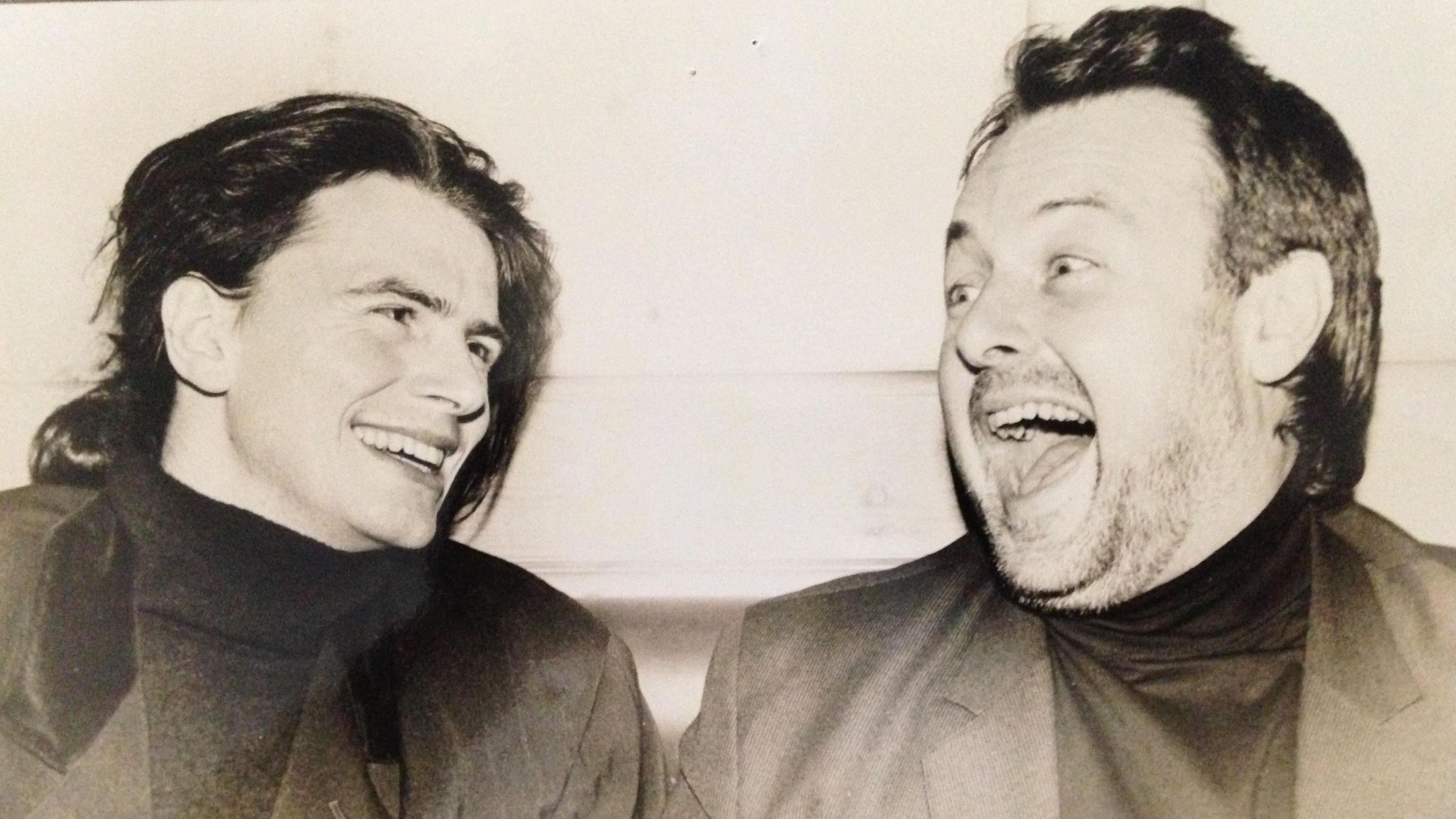 Black and white photograph shows Chris Kimsey laughing together they both wear dark polo necks and suit jackets.