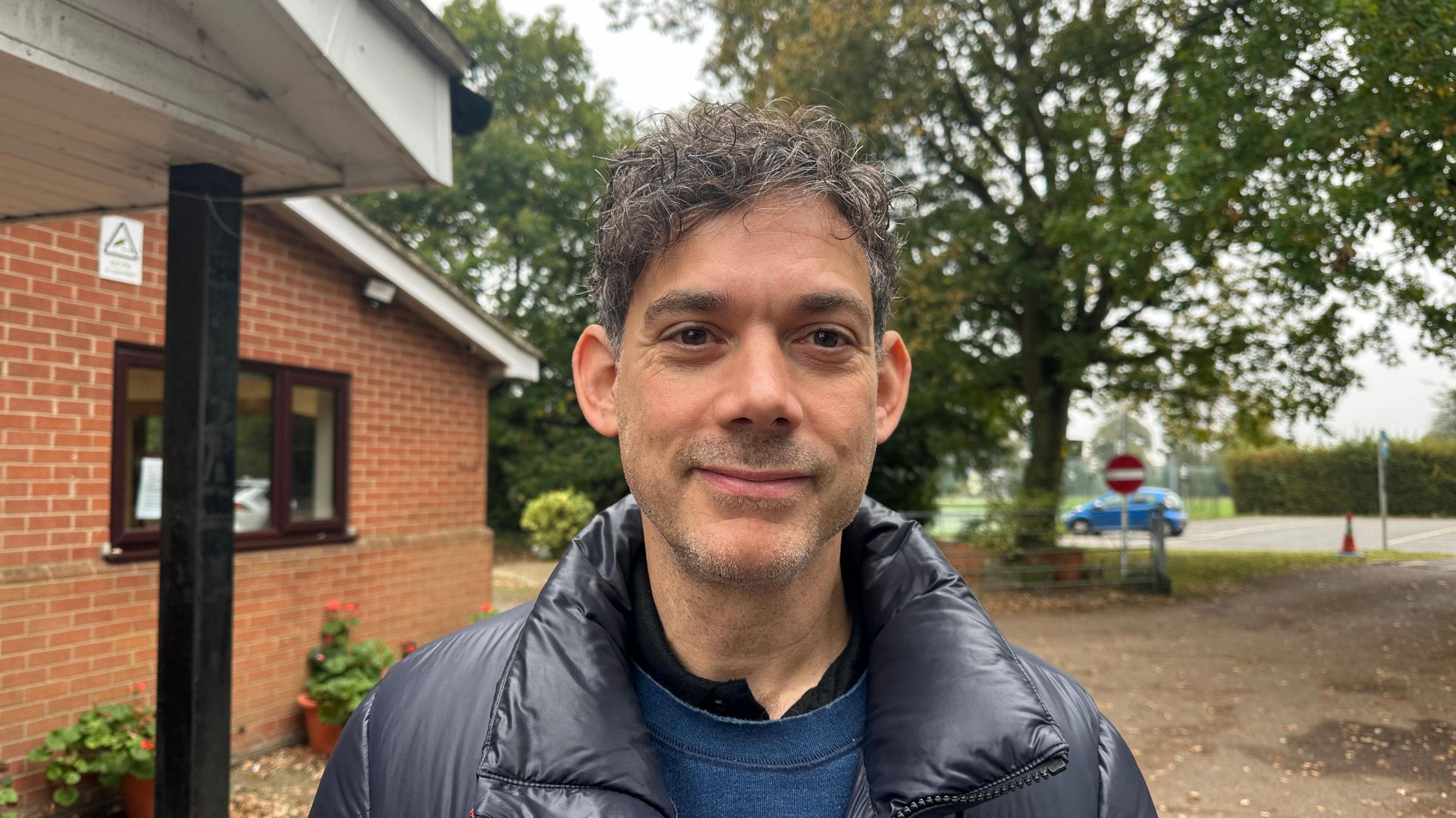 A man with grey/black hair wearing a black coat and a blue jumper smiling into the camera. 