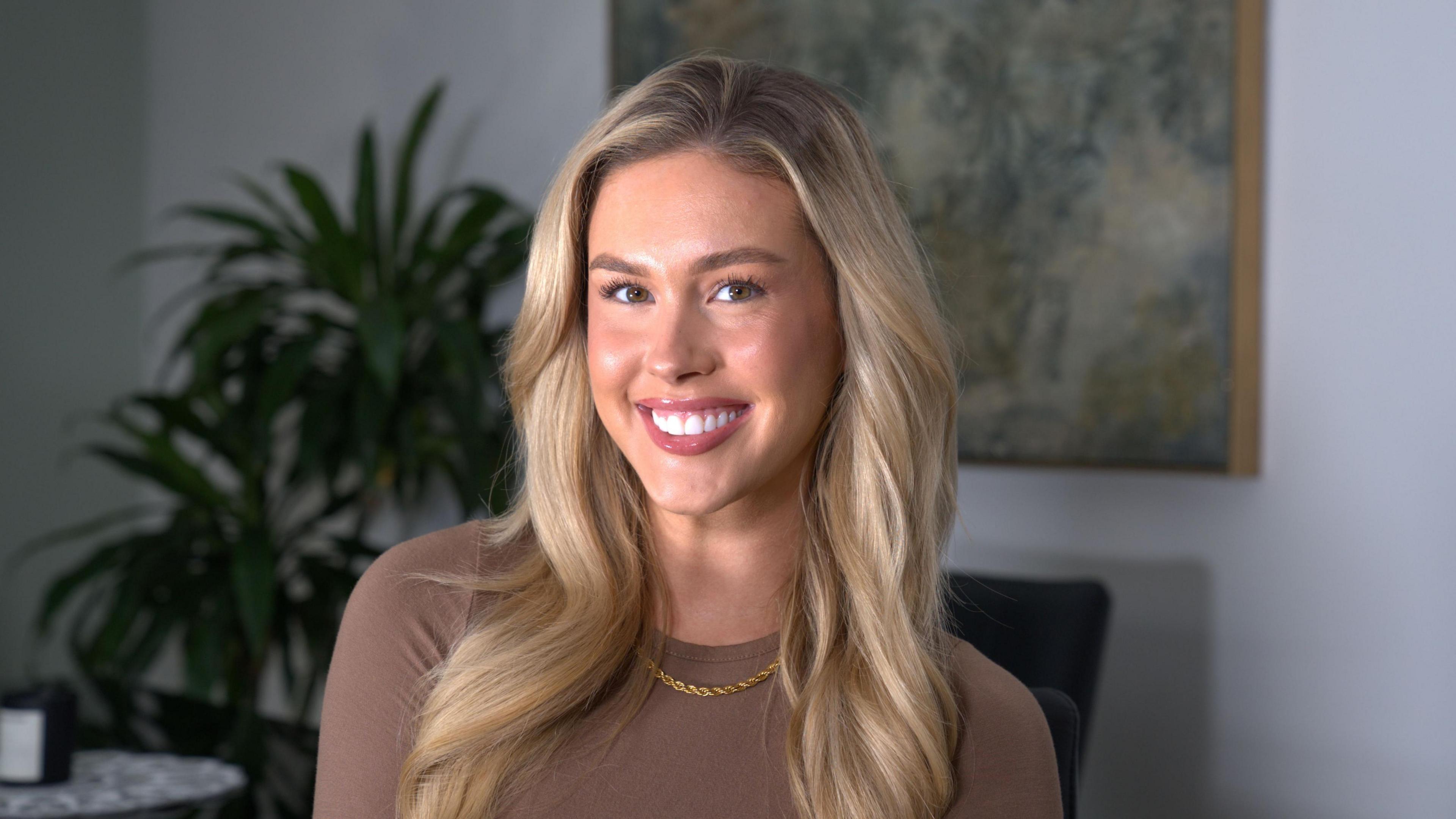 A girl with blonde hair in a brown top smiles at the camera