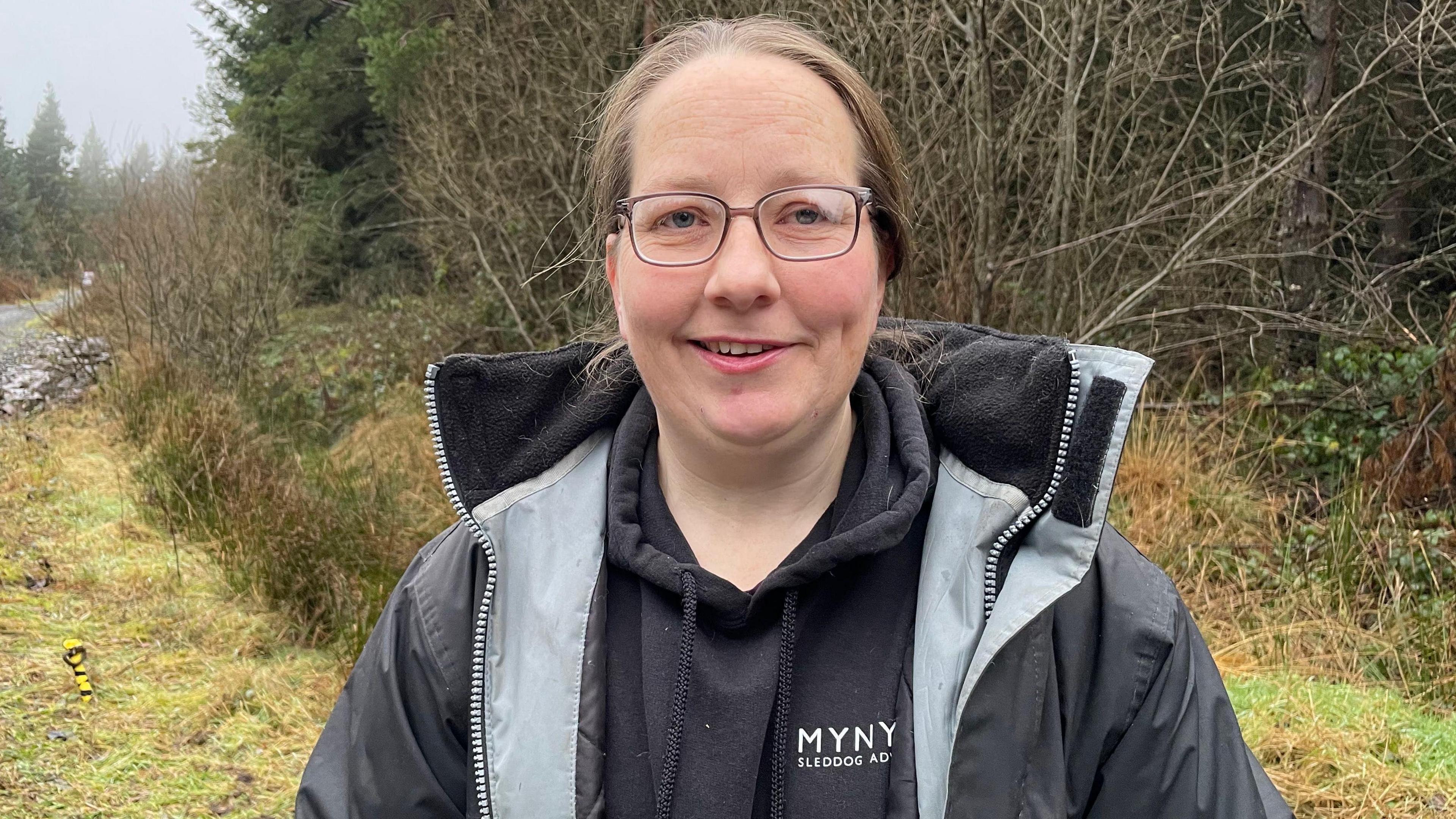 Llun o fenyw gyda gwallt brown, sbectol, siwmper du â chot du yn sefyll ar ochr llwybr coedwig