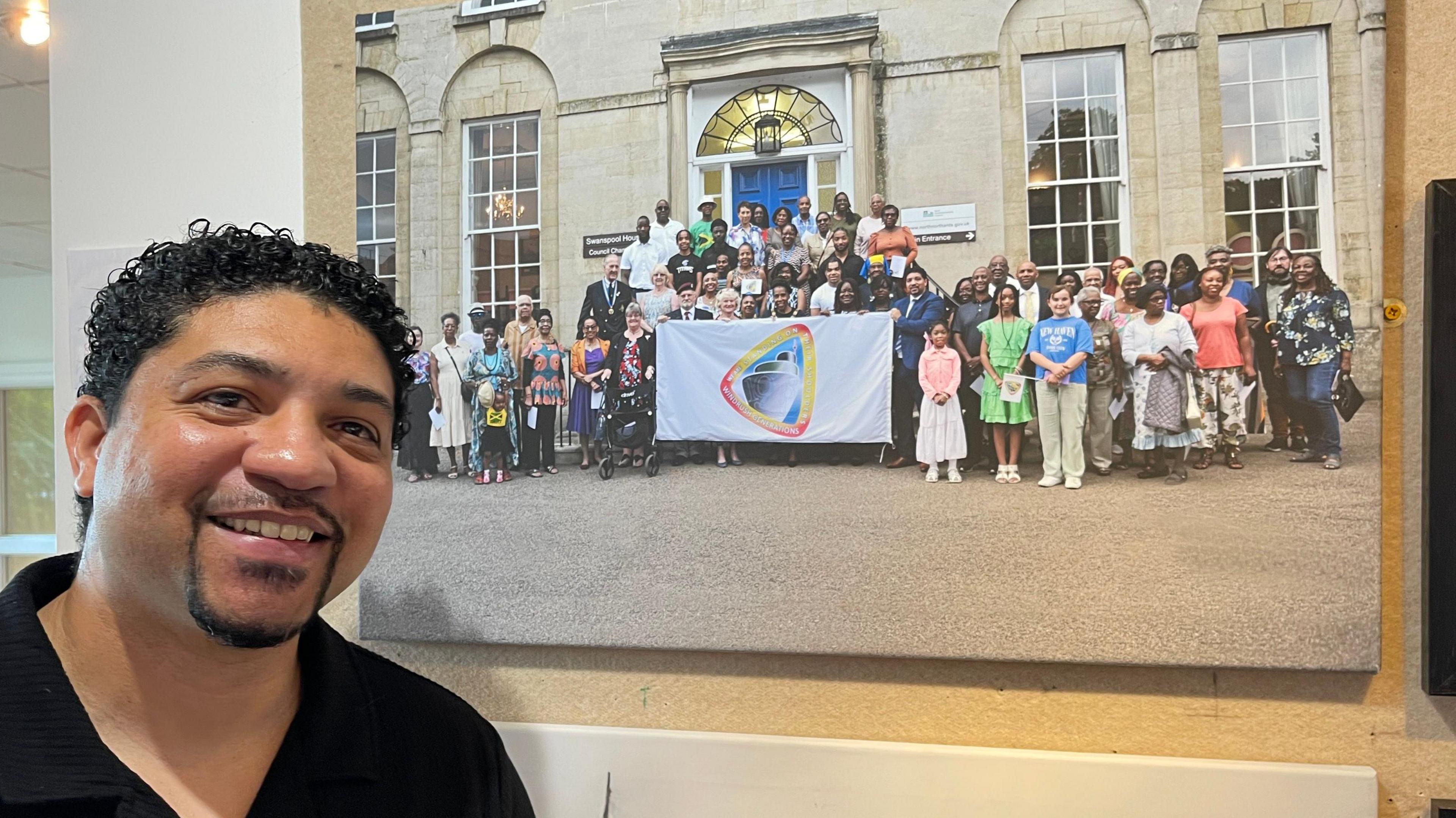 Glen Bell, who organised the exhibition, hopes it will encourage people to learn more about black history. He is smiling and has short black hair, a black goatee beard, and is wearing a black open-neck shirt. He is standing in front of a photograph of people holding a flag outside a large building.