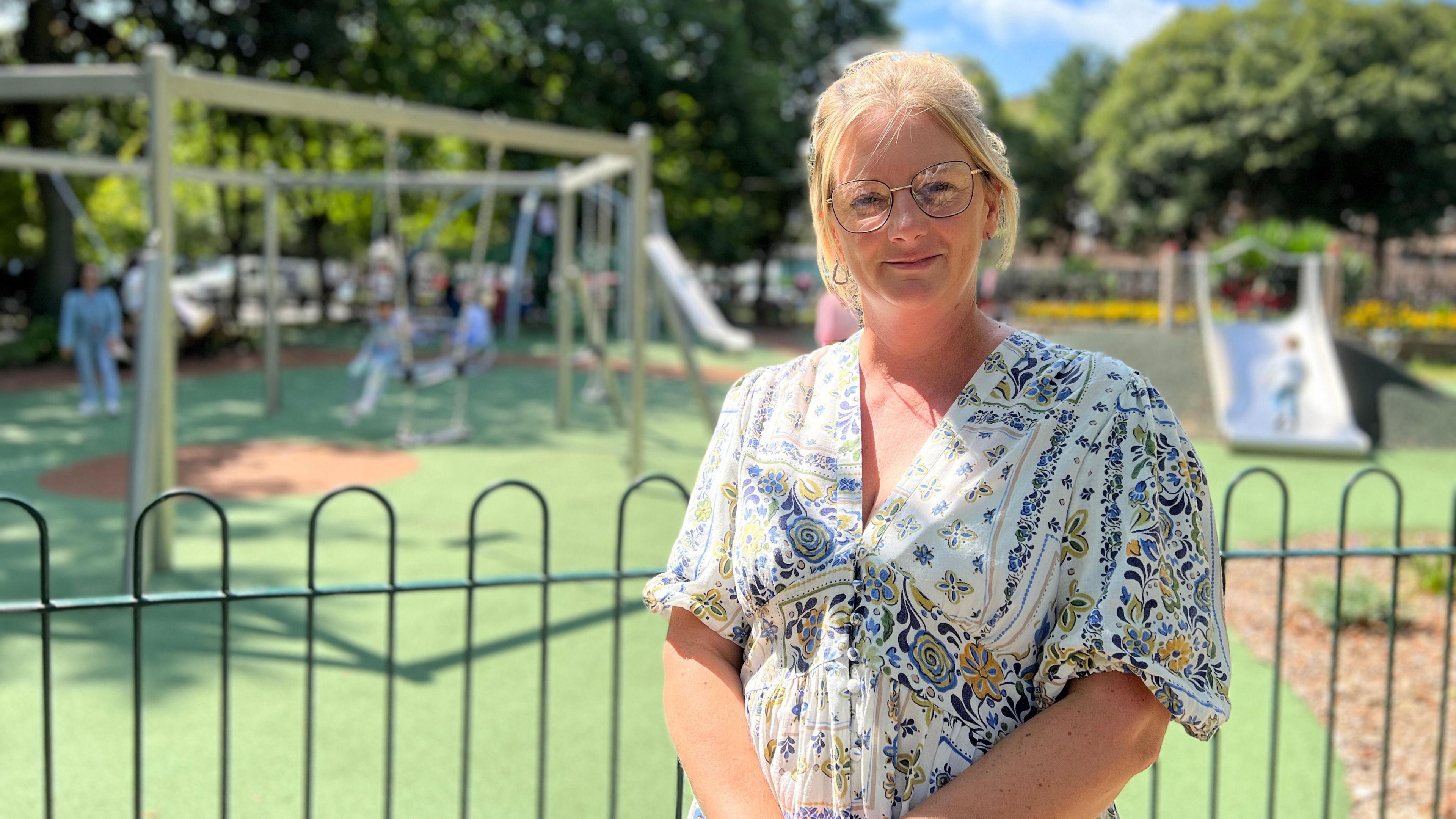 Sonja is in a floral dress stands behind a black metal fence in front of a playground with swings and slides. Children are playing in the background, and trees and a clear blue sky are visible