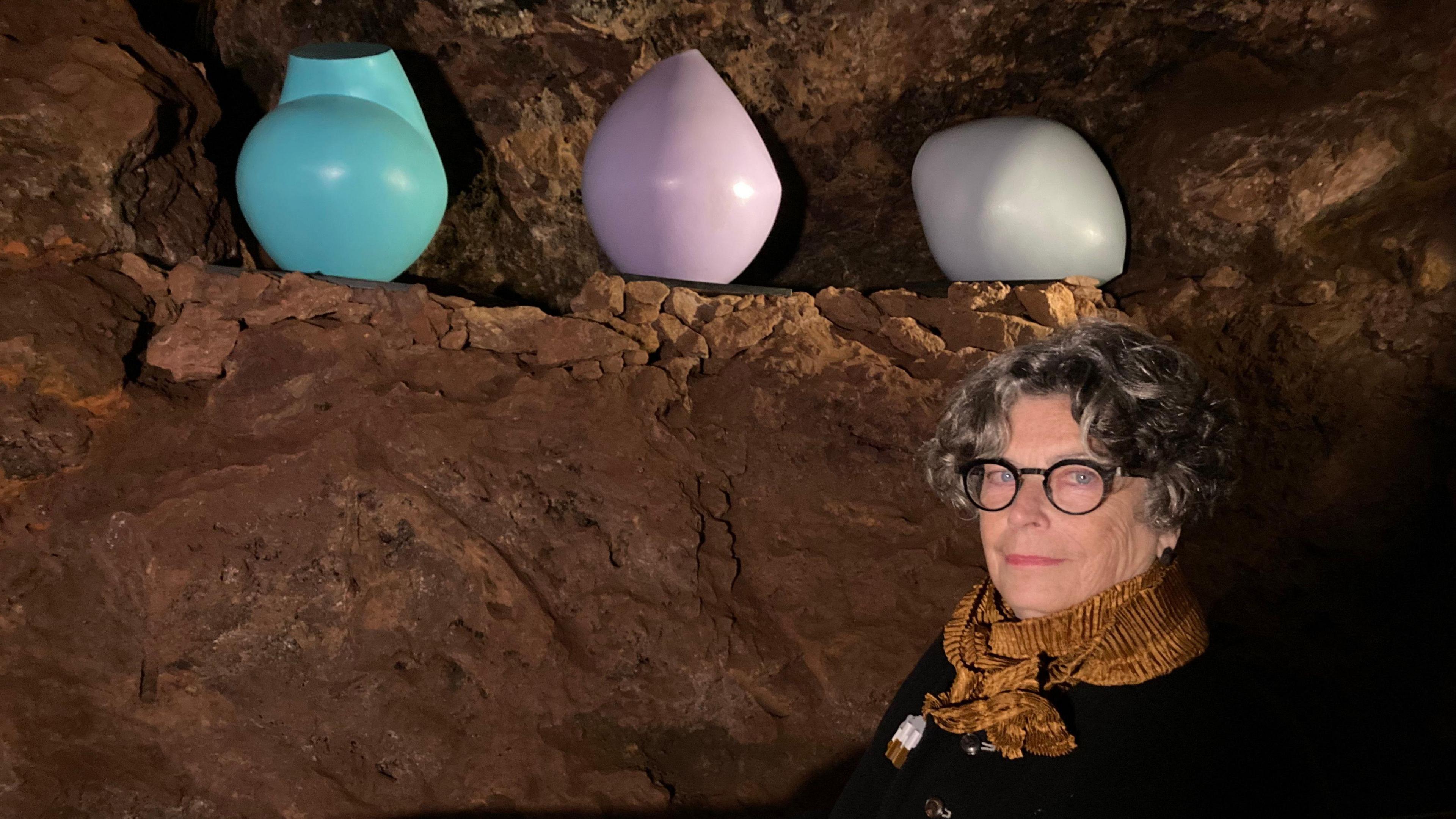 A woman with short grey curly hair, who is wearing a mustard coloured yellow scarf and black rimmed glasses, stands in a cave. On a natural ledge behind her are three small, round-ish sculptures, one is pastel blue, another is lilac, and the third is an off-white colour.
