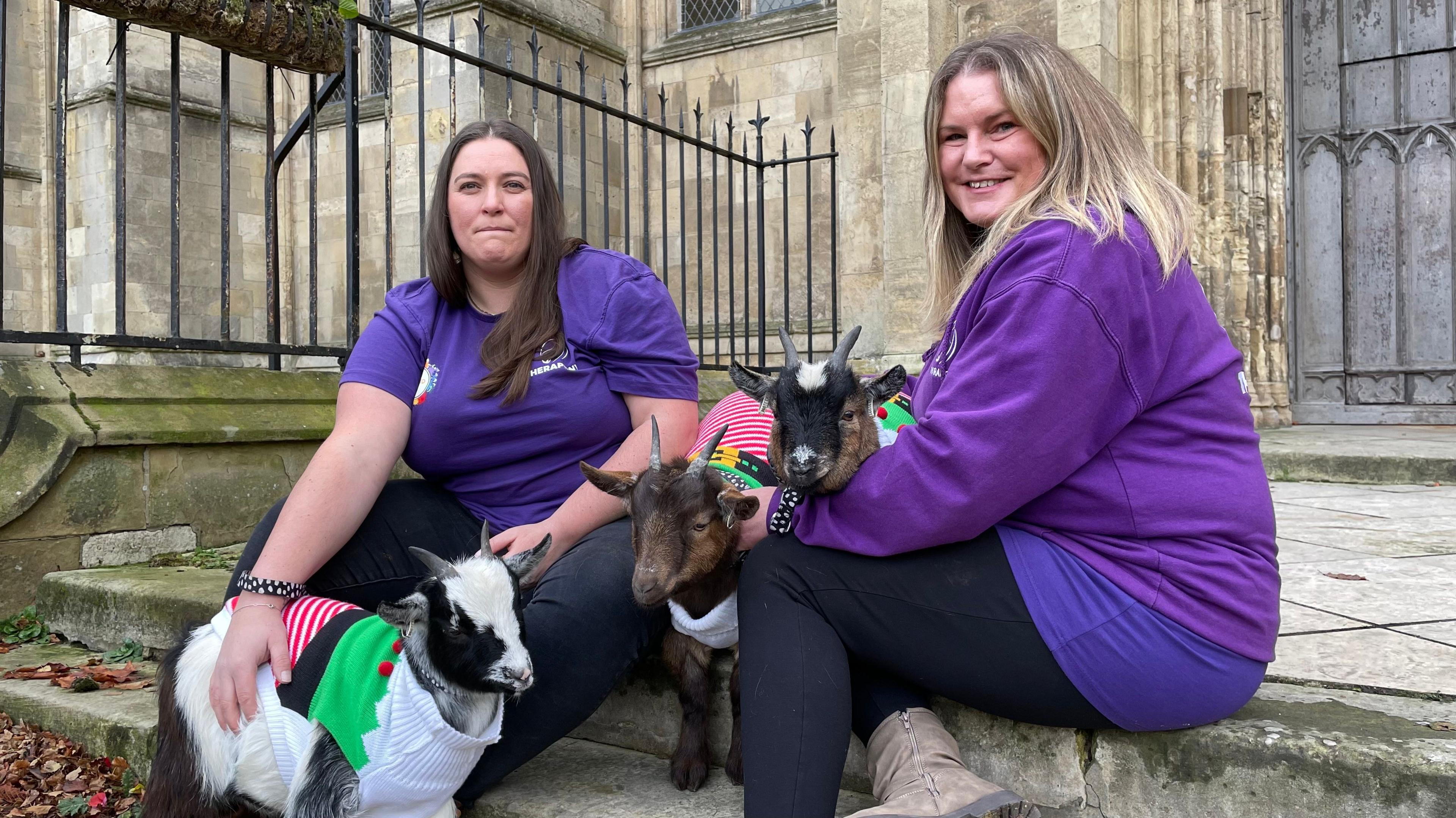 Two women, wearing purple t-shirts and black trousers sit on stone steps outside Beverley Minster. The woman on the left has long dark hair. the woman on the right has long blonde hair. They are both smiling at the camera. Between them are three miniature goats wearing Christmas animal jumpers.