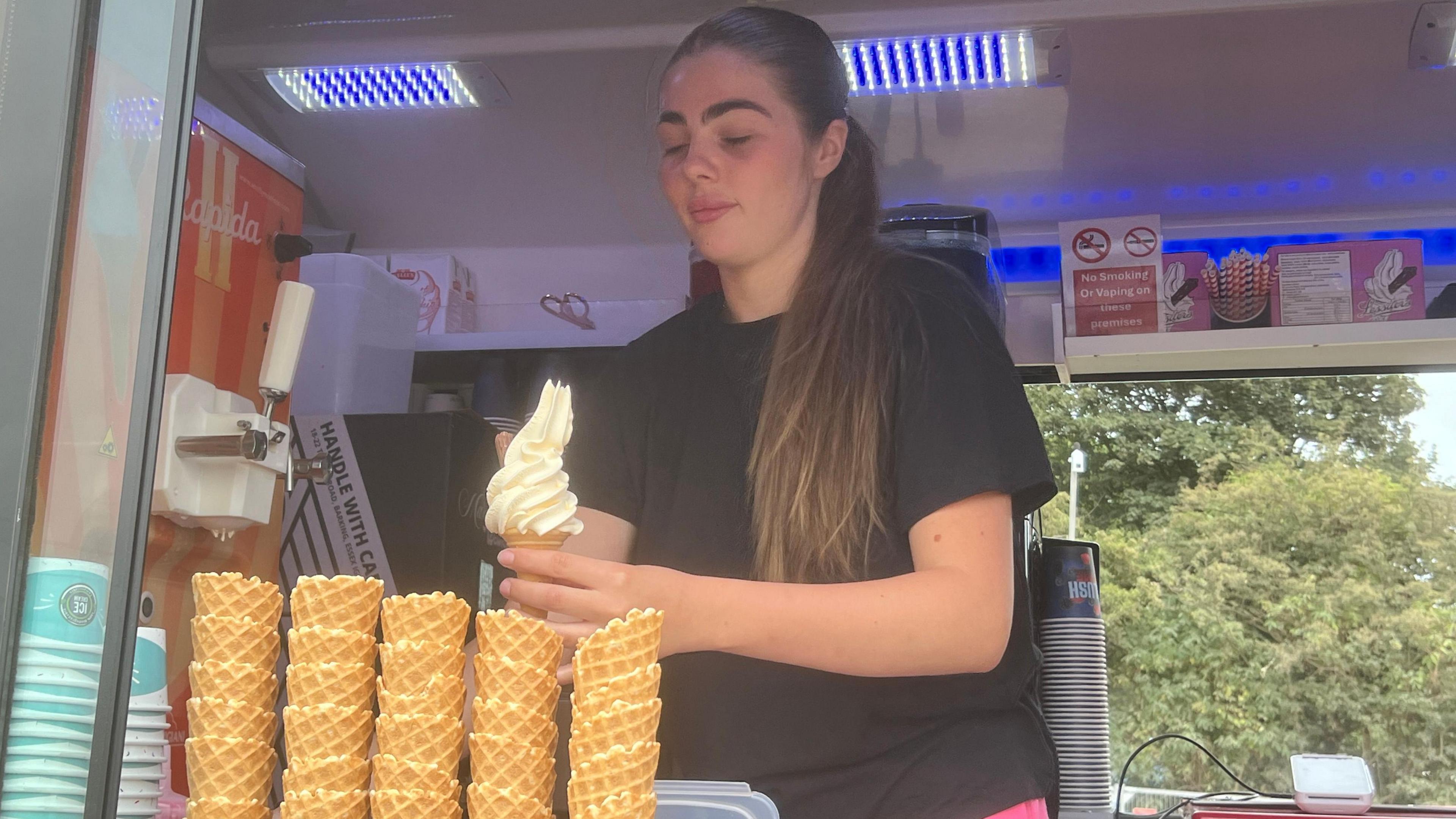 A woman is scooping soft serve onto a waffle cone. She has long hair and is wearing a black shirt.