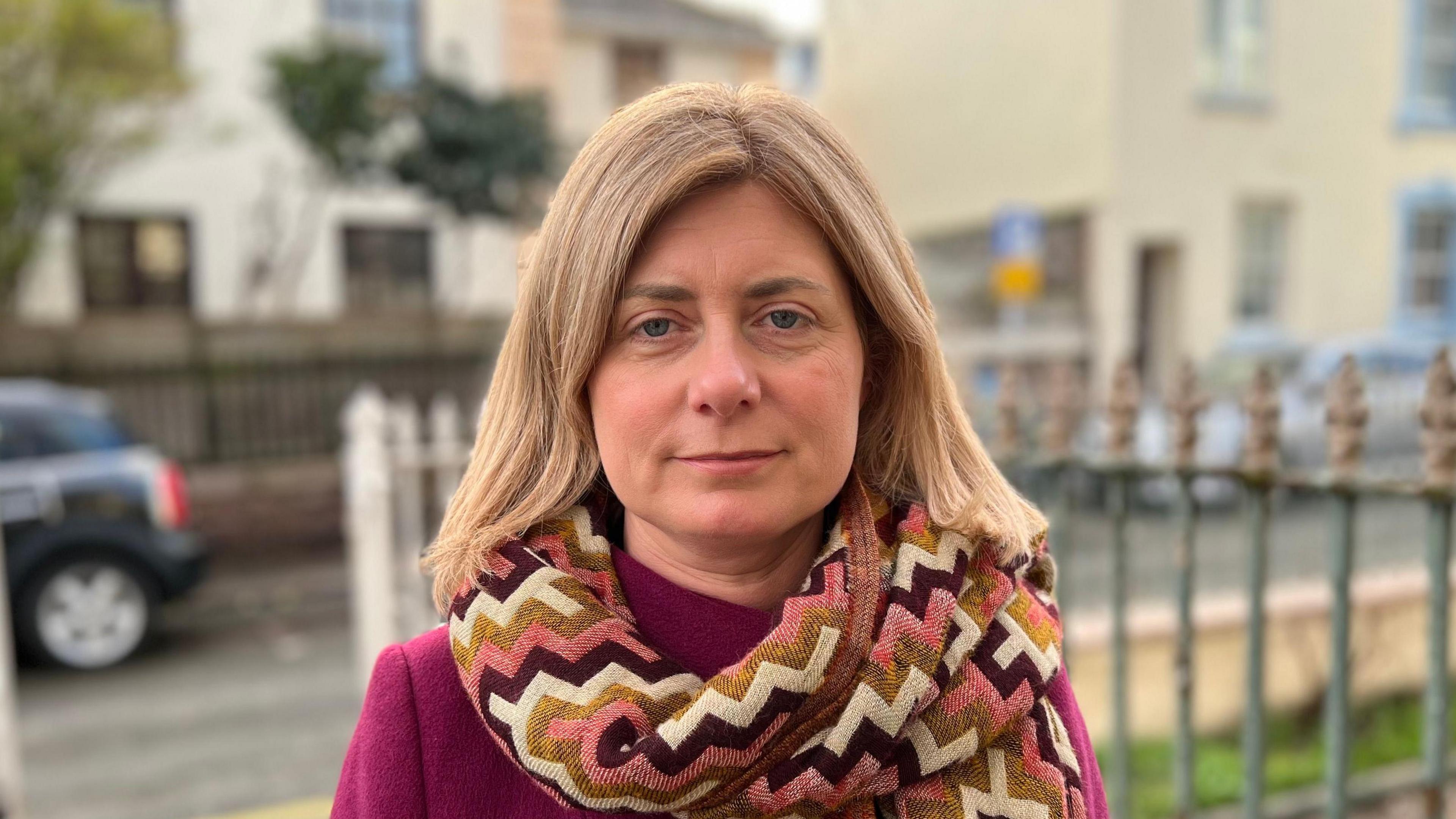 Lyndsay Feltham is a woman with blonde hair. She is looking at the camera. She is wearing a pink, cream and yellow scarf over a pink coat. She is standing in front of a street with iron railings.