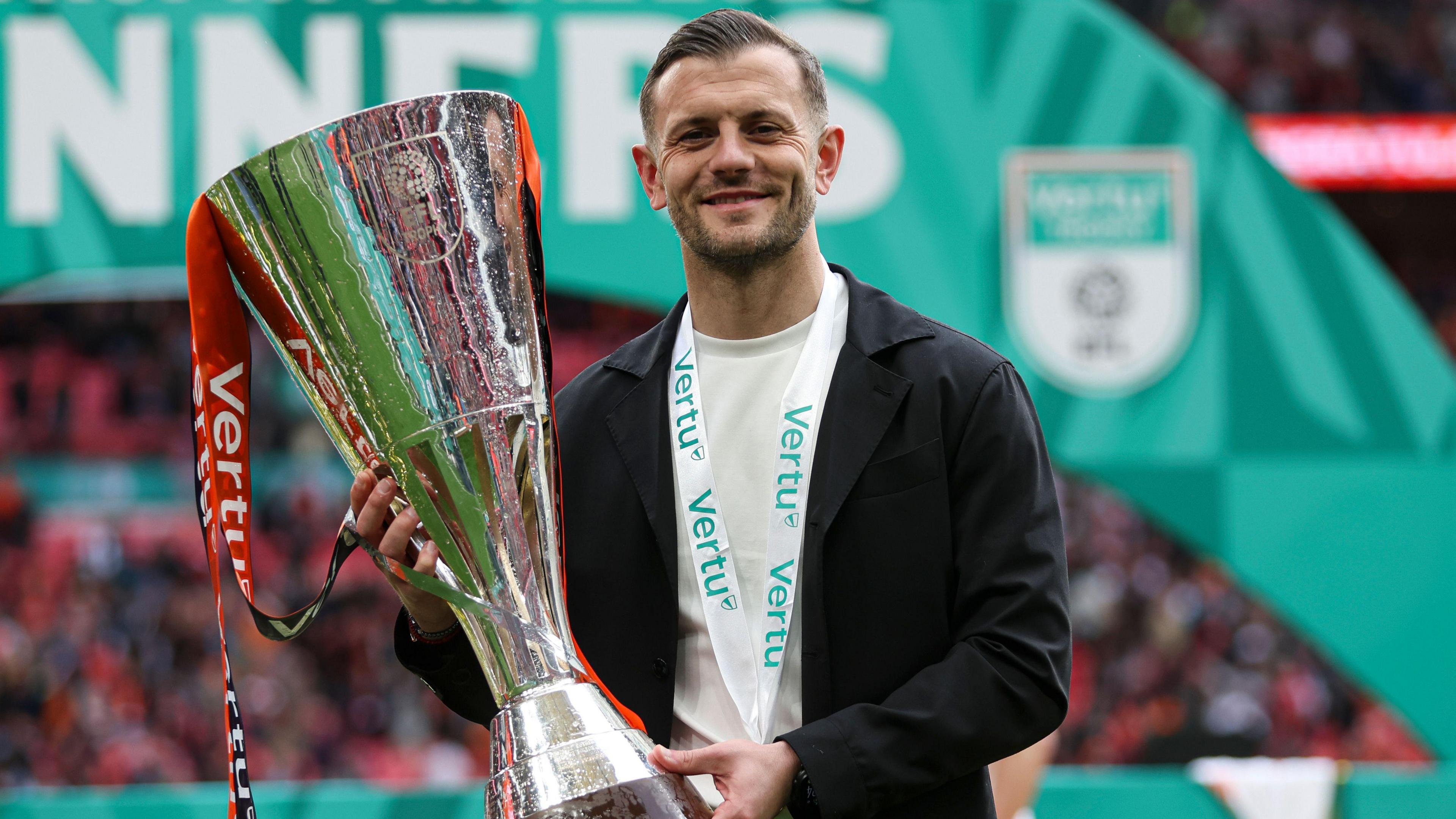 Jack Wilshere, wearing a black blazer and white shirt with a medal around his neck, smiles as he holds up the Vertu Trophy following Luton's 3-1 over Stockport at Wembley