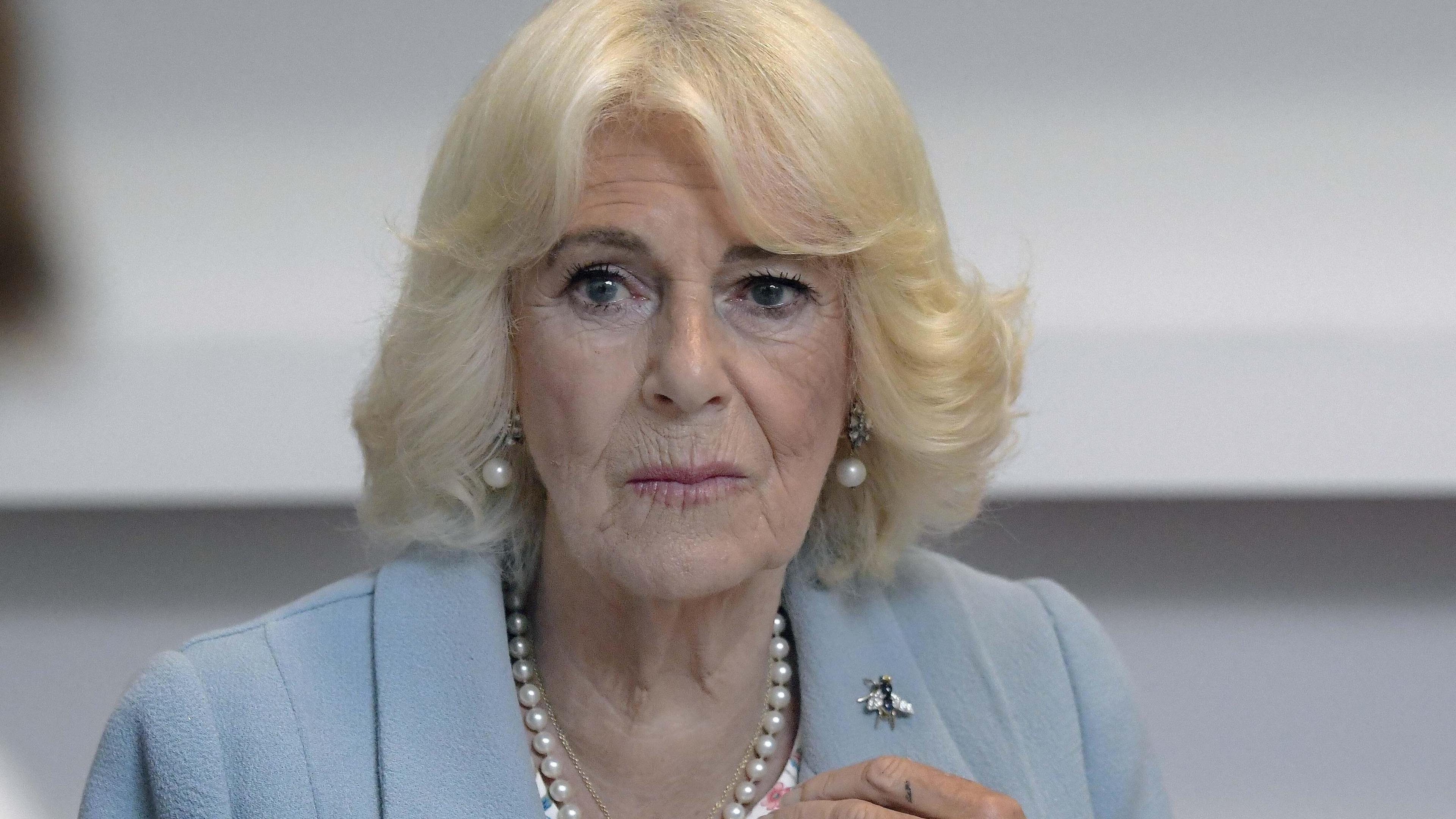 Queen Camilla wearing a blue dress and a pearl necklace and earrings looking towards the camera. 