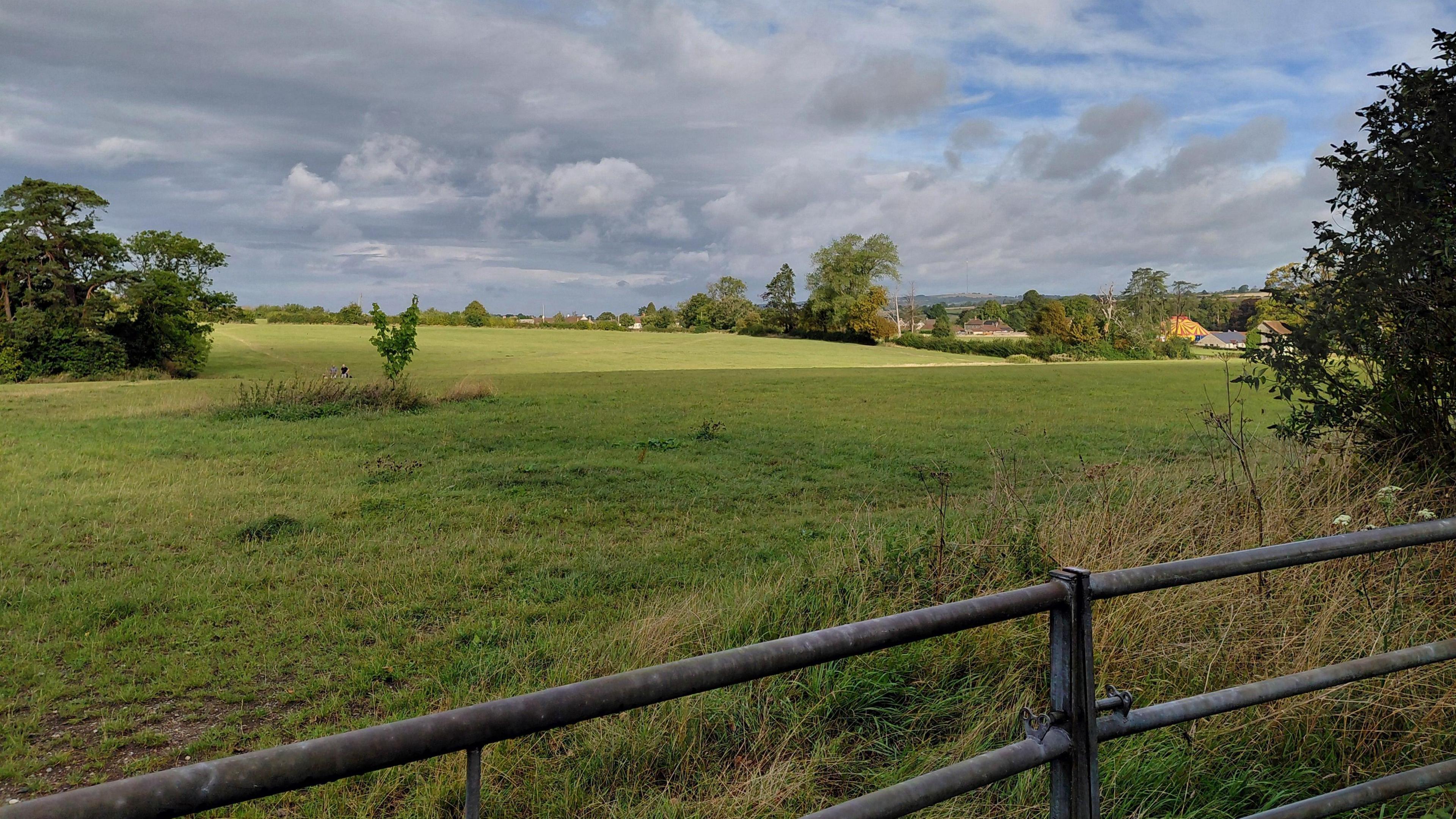 A green field behind a metal fence. There are some clouds in the sky but the sun is shining through.
