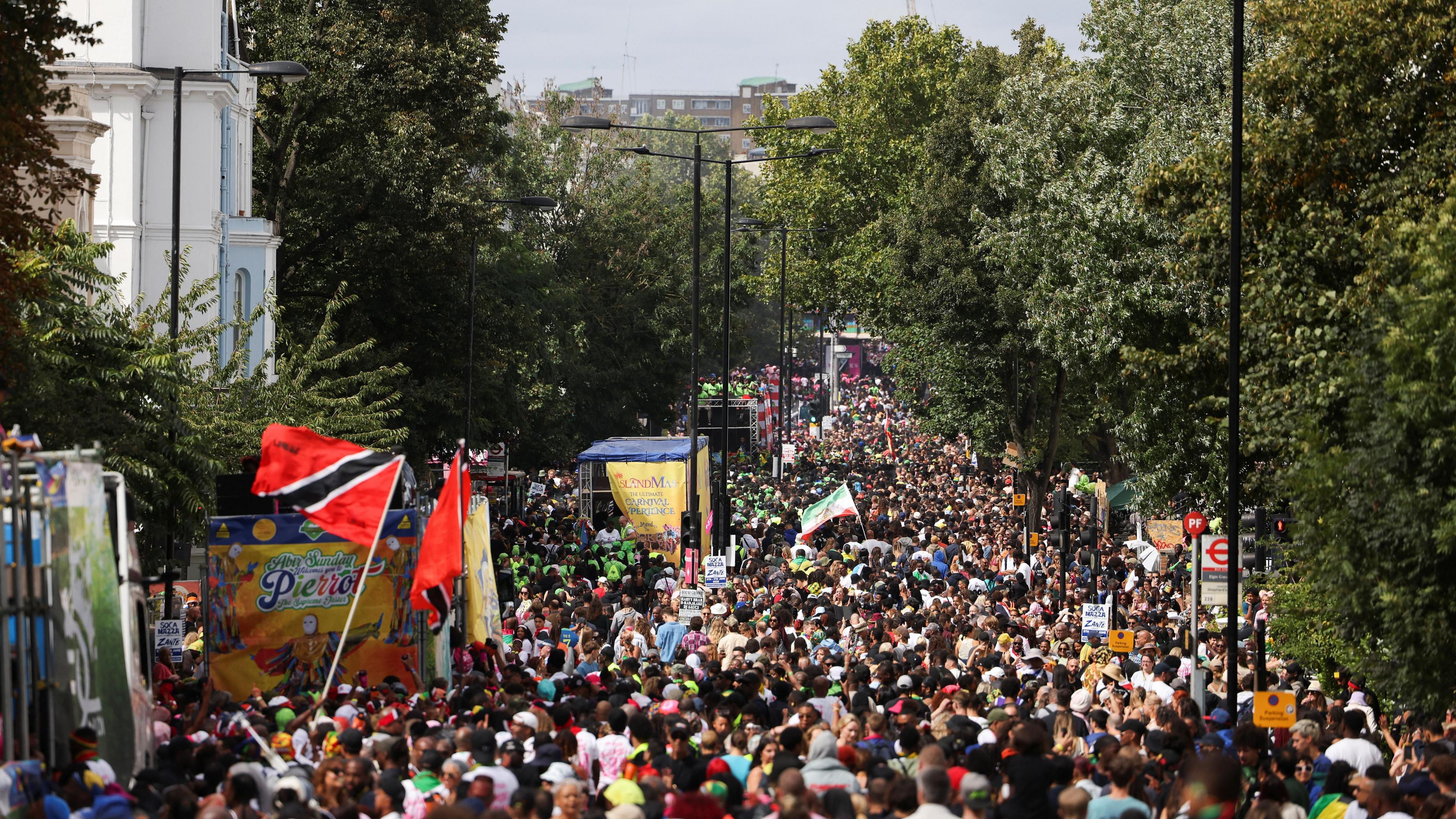 A large crowd fills a tree-lined street during Notting Hill Carnival, with colourful floats, flags - including the Trinidad and Tobago flag - and banners visible among the dense procession.