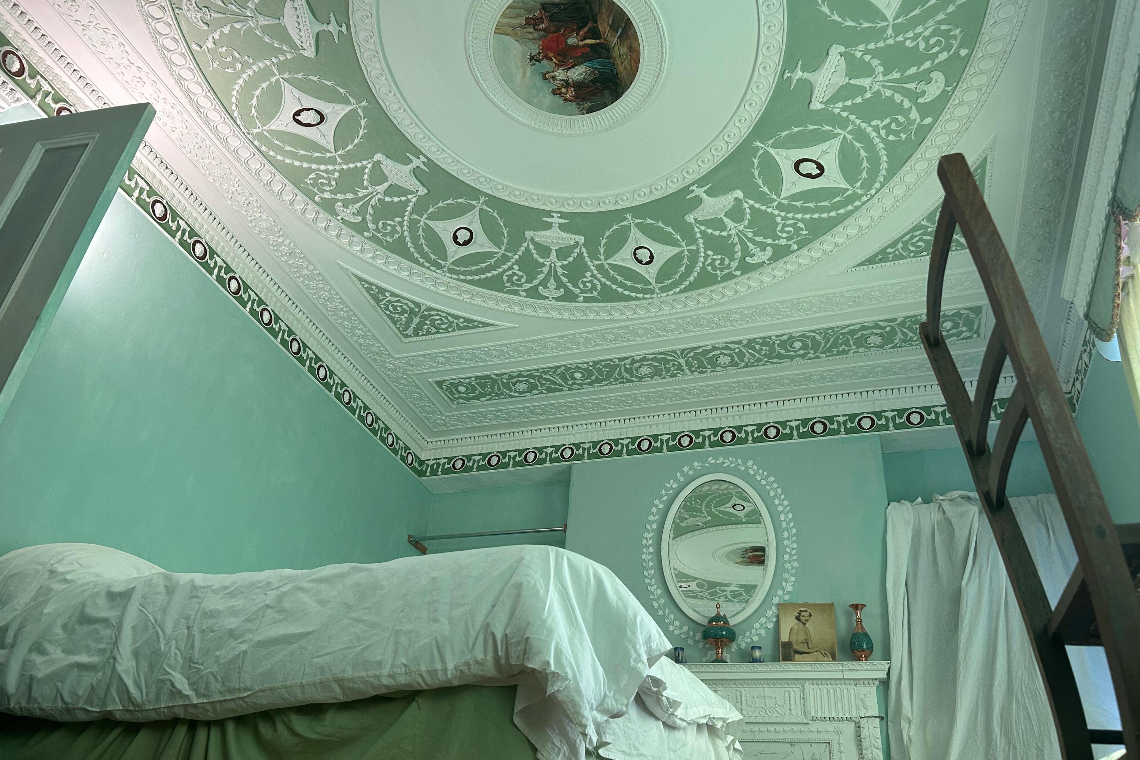 A wide image of one of the decorated bedrooms. The ceiling is painted a pastel green colour with intricate white neoclassical detailing, including a circular piece of artwork in the middle depicting a group a people, and concentric circles going out from it with more circular patterns within one band. A dark green funky border surrounds the room. In the middle of the room there is a double bed with white bedding. Behind the bed, which is in the foreground of the image, there is a white mantel piece with a oval mirror above. There are several trinkets on the mantel piece and there is a wooden chair in the bottom right of the image.