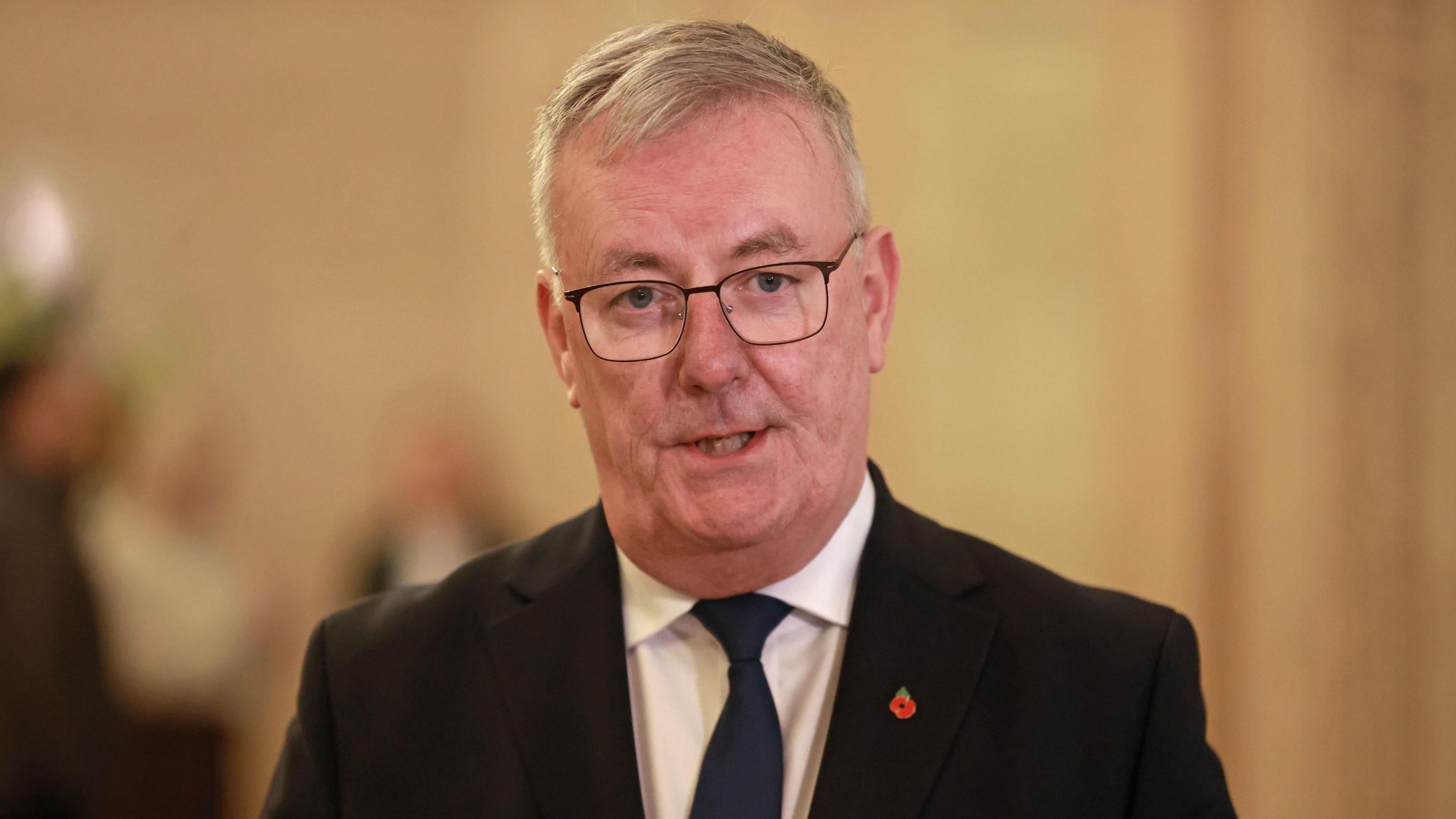 Mike Nesbitt - a man with grey hair wears square framed glasses as well as a poppy pin, dark suit jacket, a white collared shirt and dark tie is speaking.