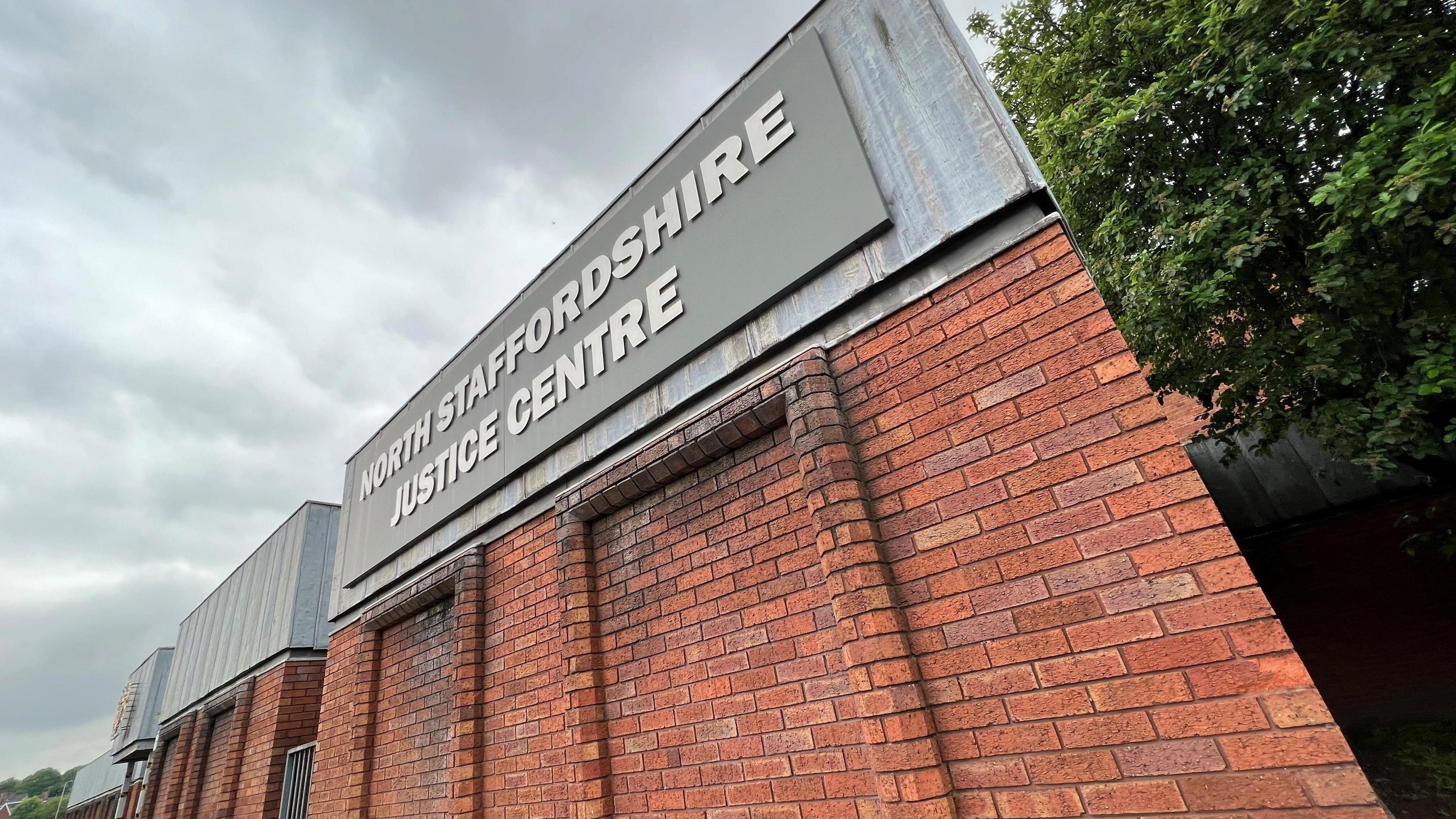 A red brick building, viewed from a corner, with metal flashing around the top. There is a grey sign with lettering that reads North Staffordshire Justice Centre.