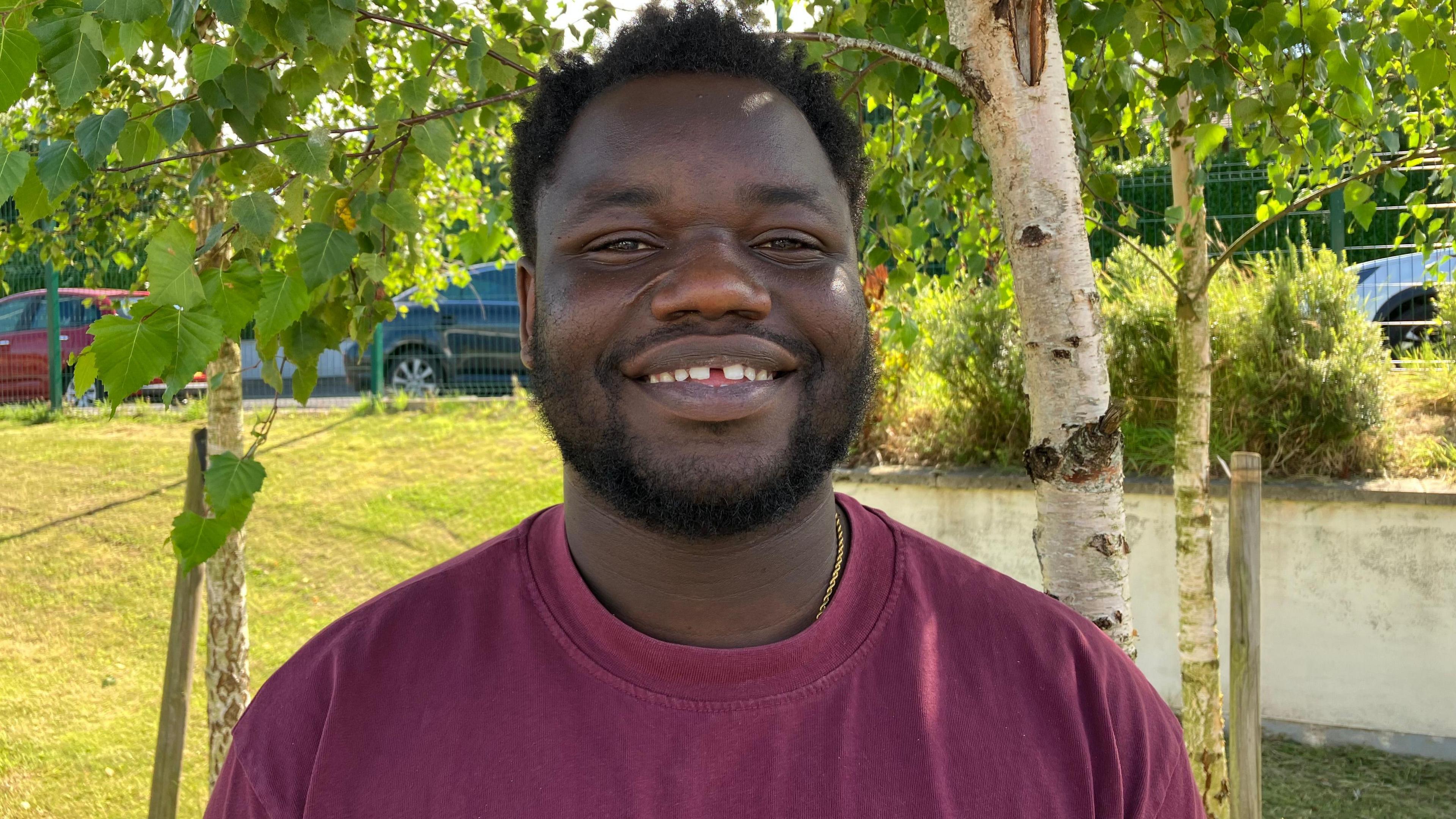 A man, smiling at the camera, wearing a burgundy tshirt. There is grass and trees behind him.