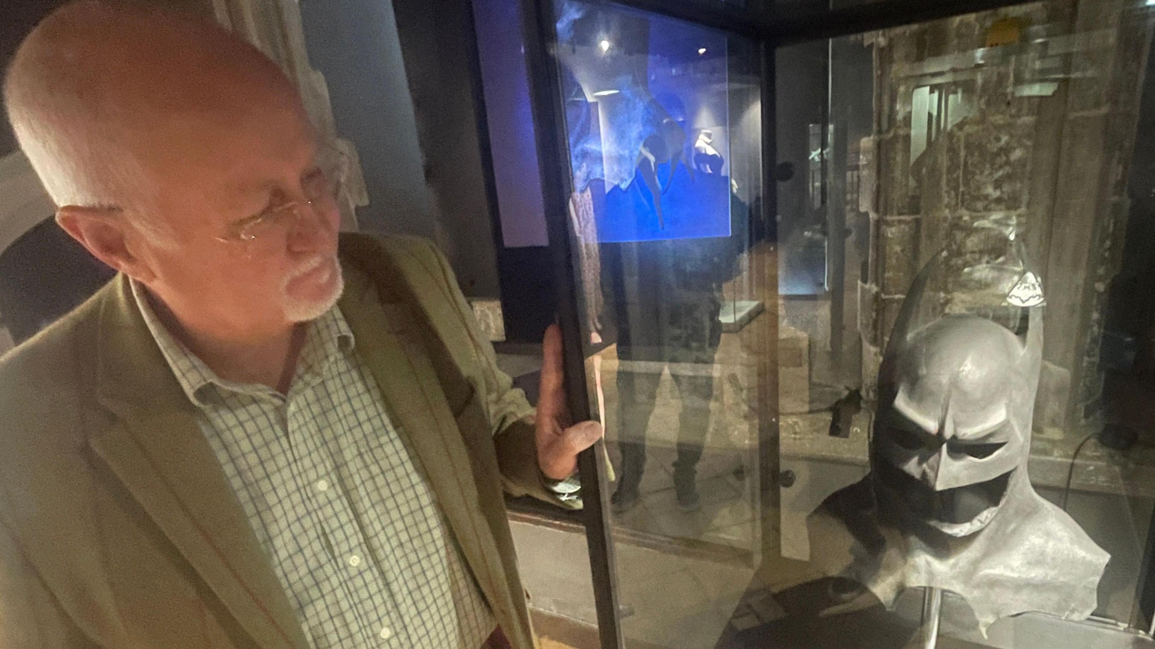 Stephen Rivers, director of Kudos Memorabilia, standing next to a glass cabinet in which there is a Batman cowl on a stand. Mr Rivers is wearing  green blazer over a shirt and glasses. He is looking at the cowl.