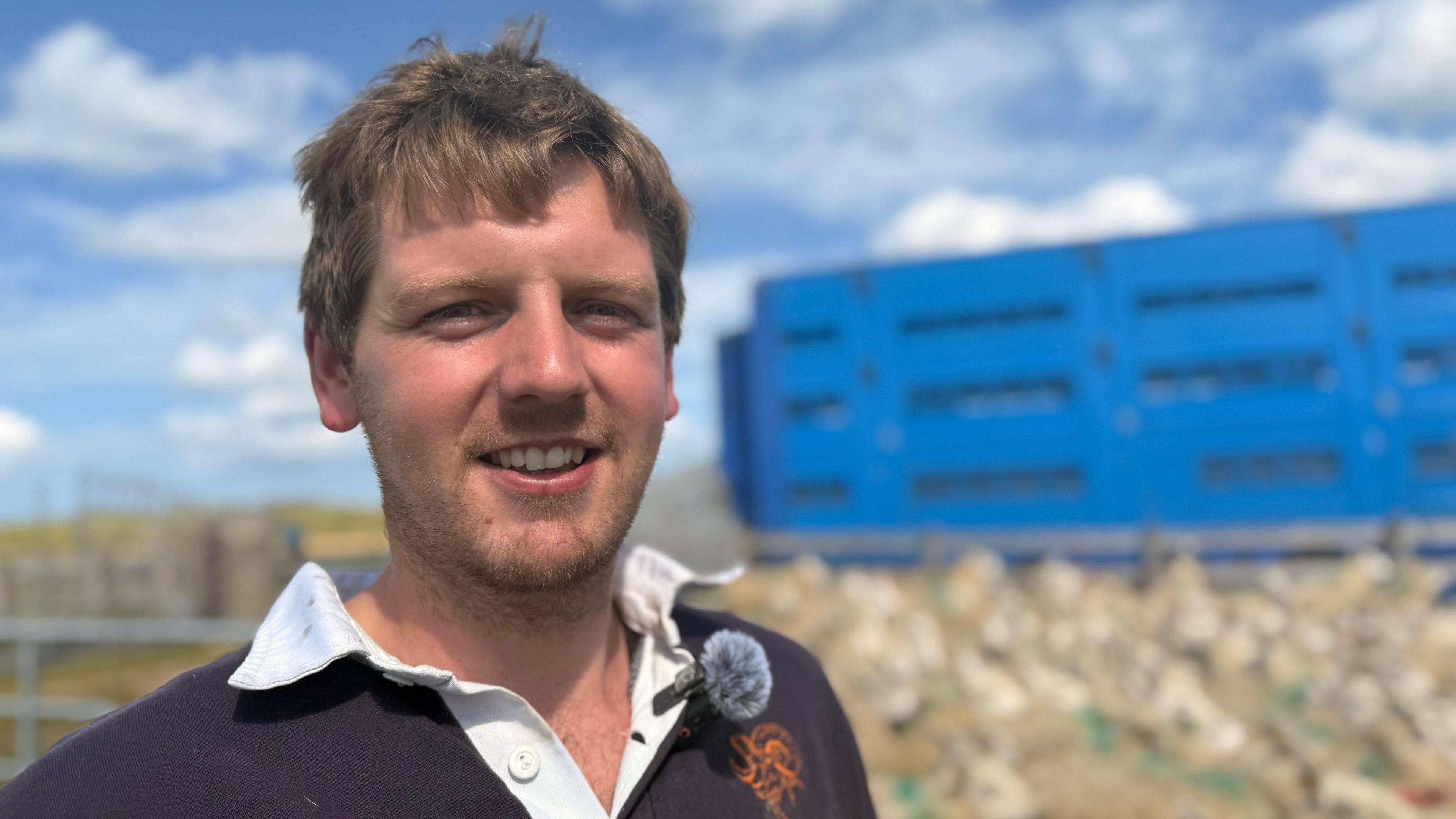 Aaron with short brown hair smiling at camera. He is wearing a navy jumper with a white collar. He has a small microphone with a grey fury wind shield on his collar. The background is blurred and has sheep and a blue transport lorry