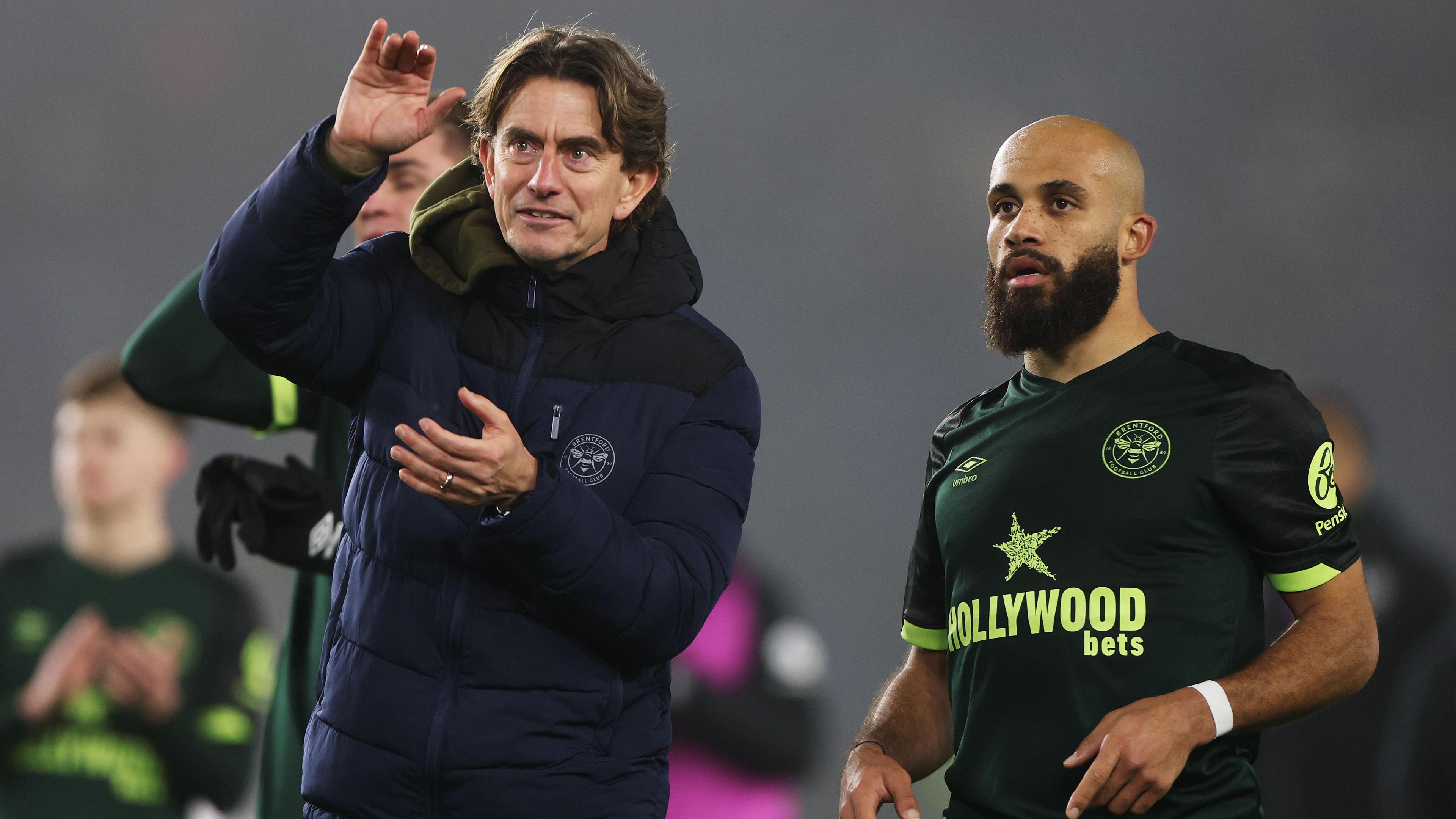 Brentford manager Thomas Frank wears a long blue padded jacket and uses his hands to emphasise a point to Bryan Mbeumo who stands next to him wearing a green Brentford away shirt. Other blurry Brentford players can be seen applauding in the background