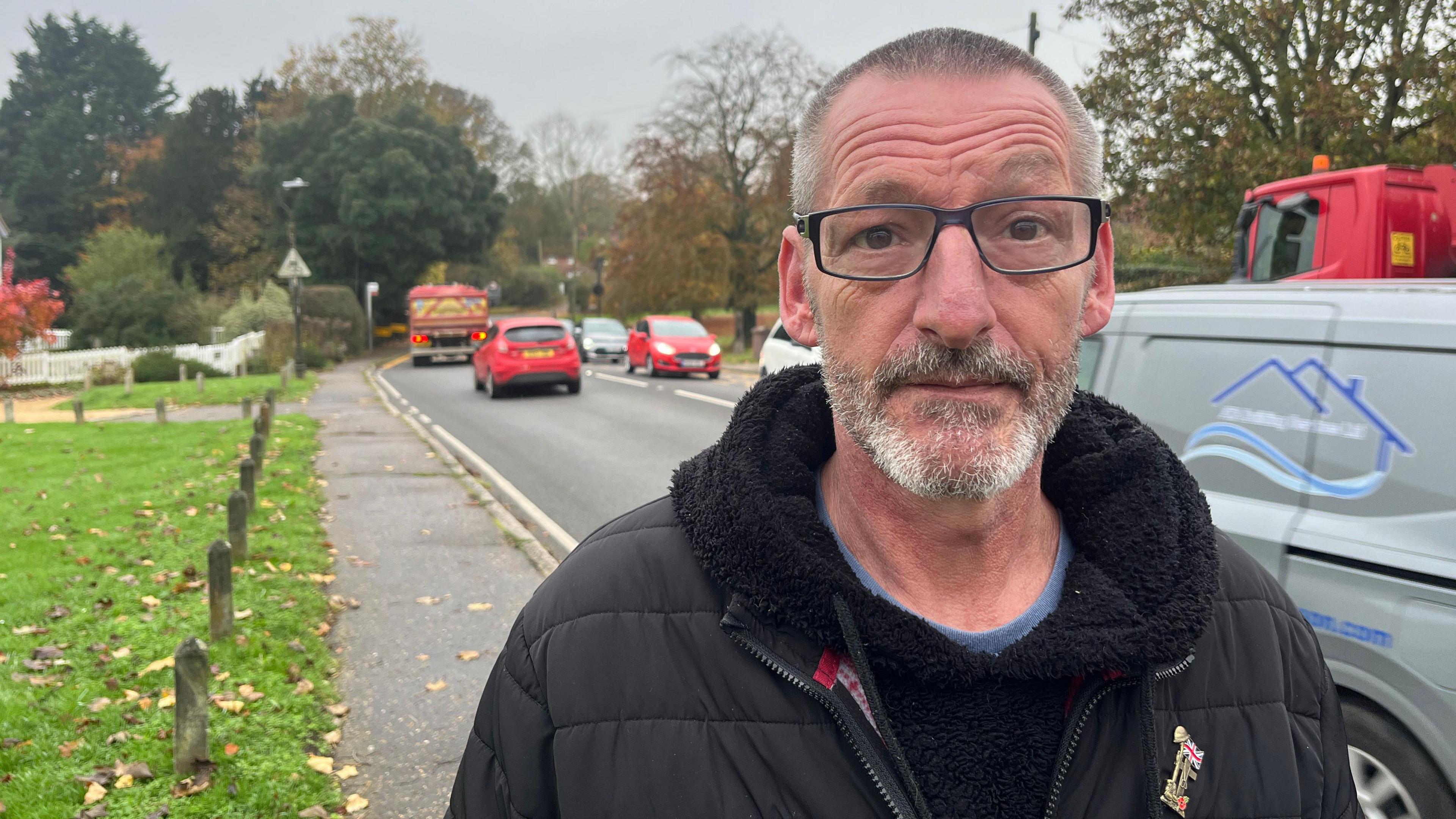 Jason Coleman, who is wearing a black jacket with a black hoodie underneath, looking at the camera expressionless. He has short grey hair. He is standing on the pavement next to Stock Road on a cloudy day. Cars are travelling on both sides of the road, which is lined with grass and trees.