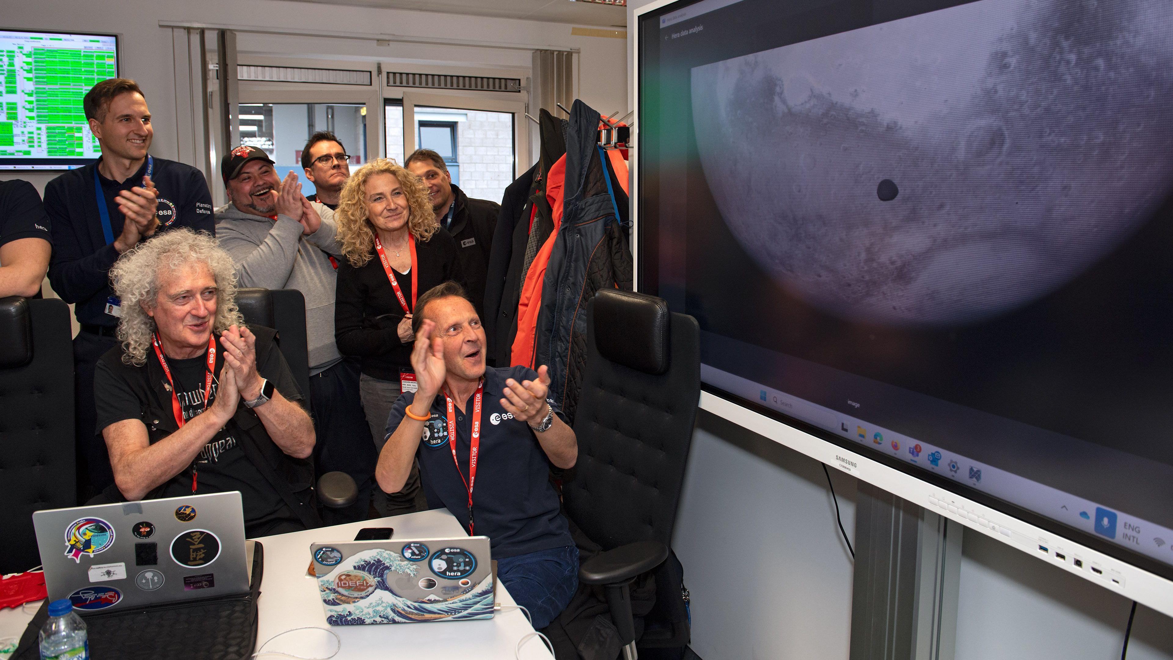 A group of scientists applaud at an image of the moon Deimos, including Sir Brian May, with long white hair