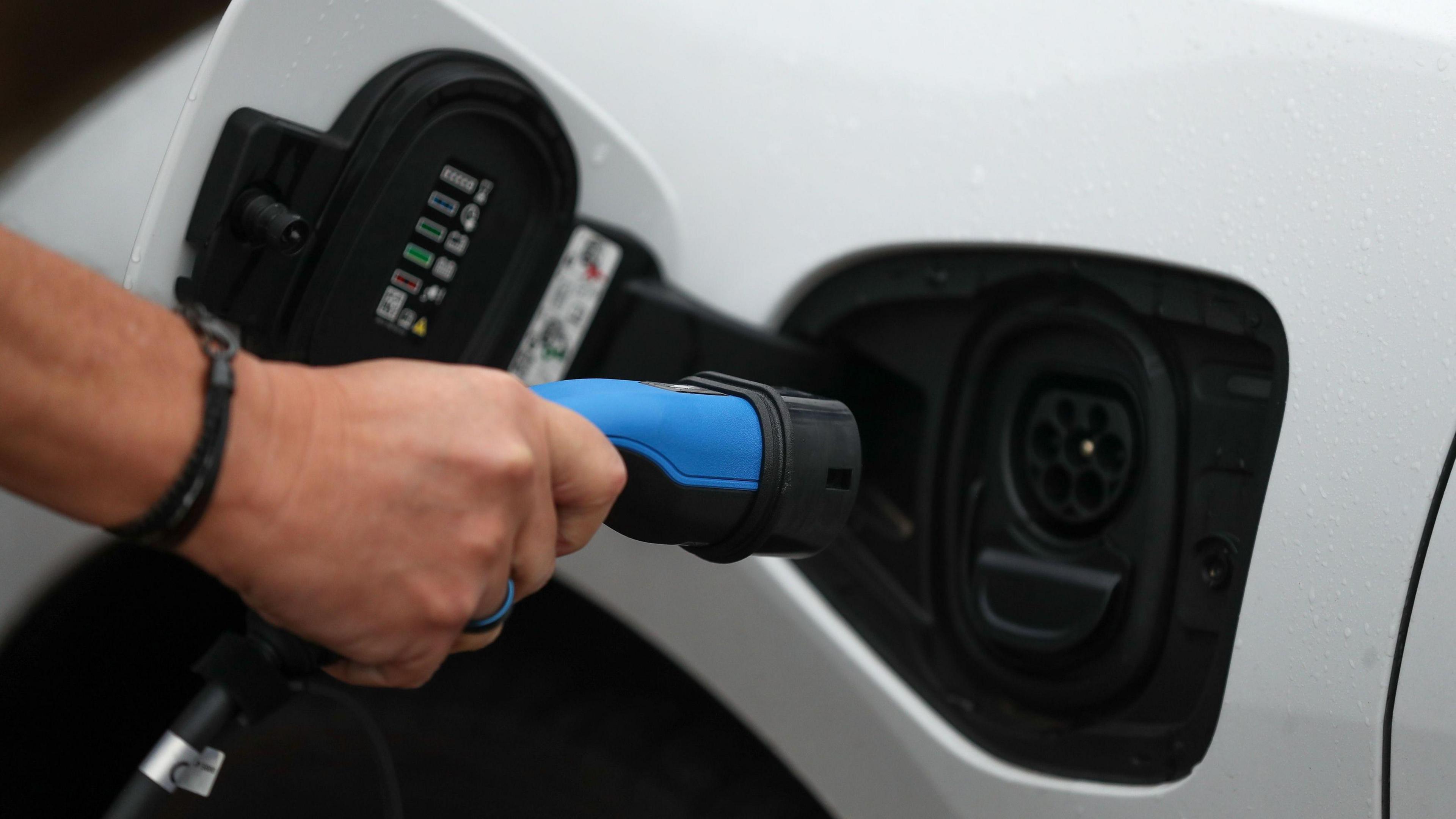 A picture of someone holding a charger. They are wearing a black wristband and holding a blue charger. It is a white vehicle with a black port.