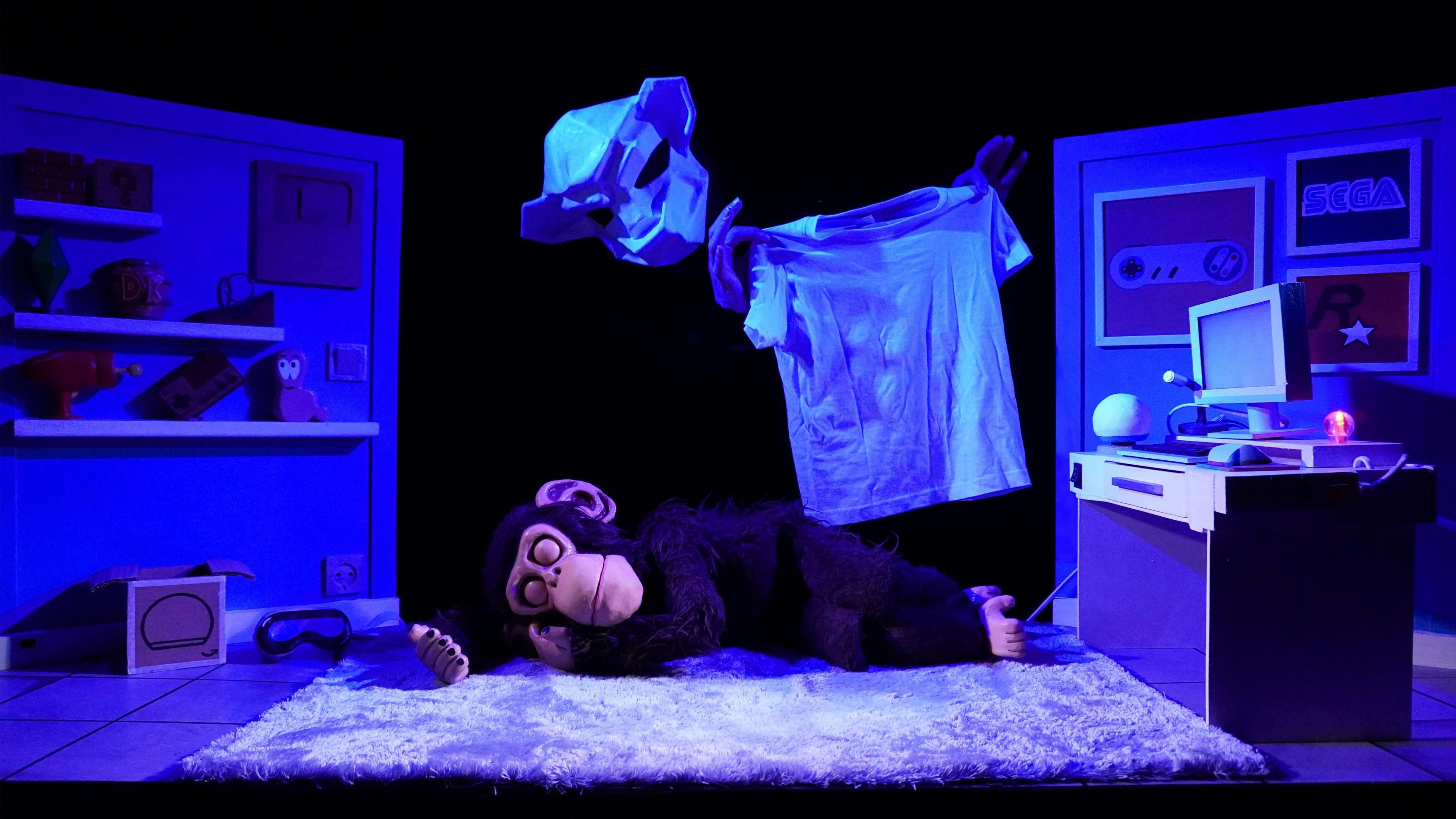 A puppet monkey lies on a stage, set up to look like a bedroom, with a computer, posters on the wall and shelves. Above the monkey, a ghostly monkey puppet hovers, holding a white T-shirt.