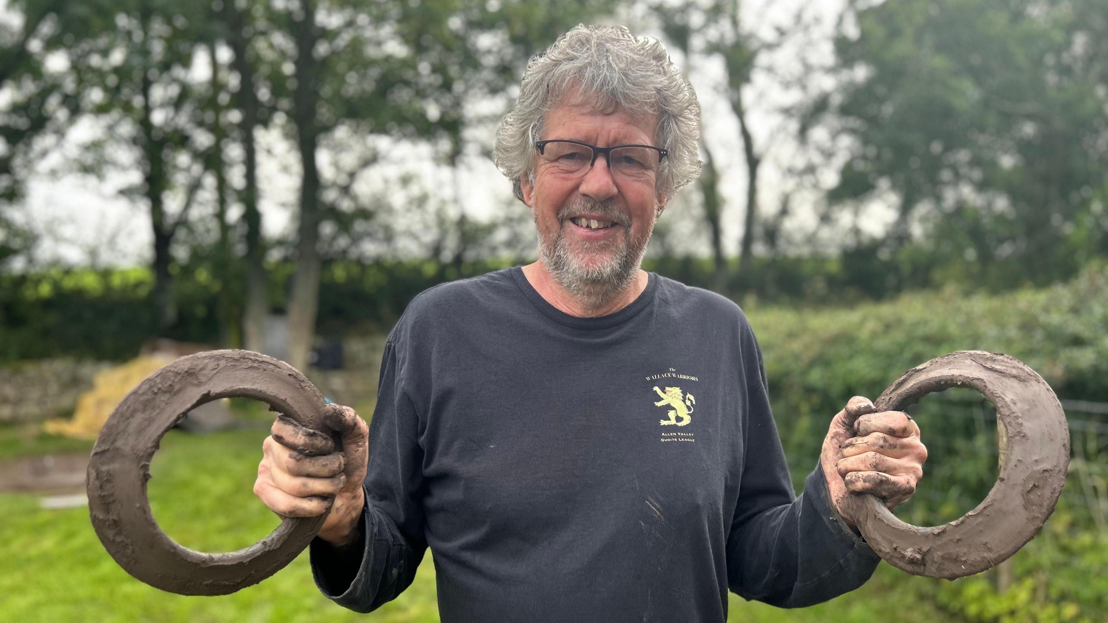 Richard Macdonald is smiling and holding a quoit in each hand. He has wavy grey hair and is wearing a black top and glasses. He is standing on grass and there are blurred trees in the background.
