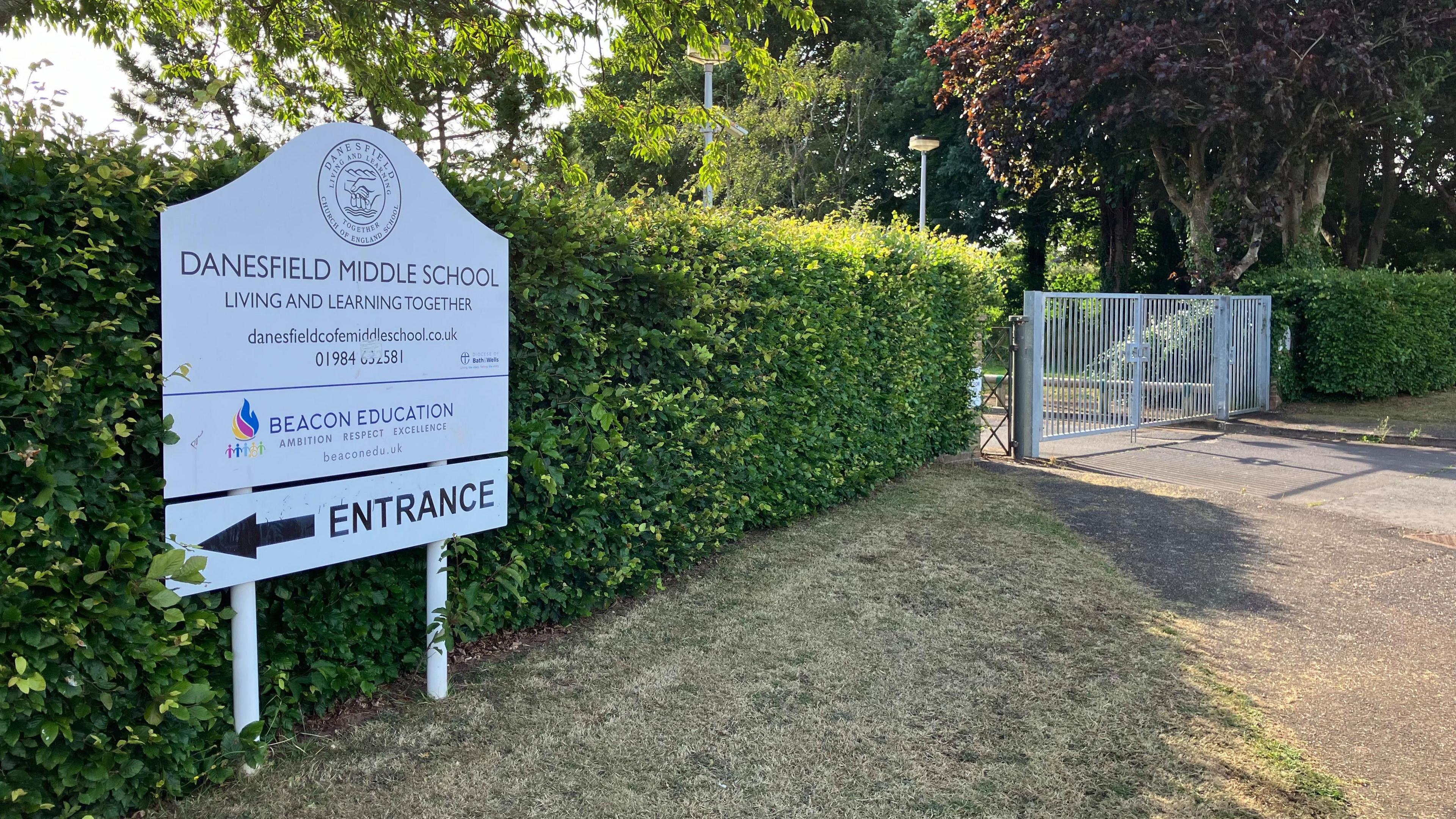 A view of a school building. The sign in front says Danesfield Middle School. There is a green hedge and a school gate.