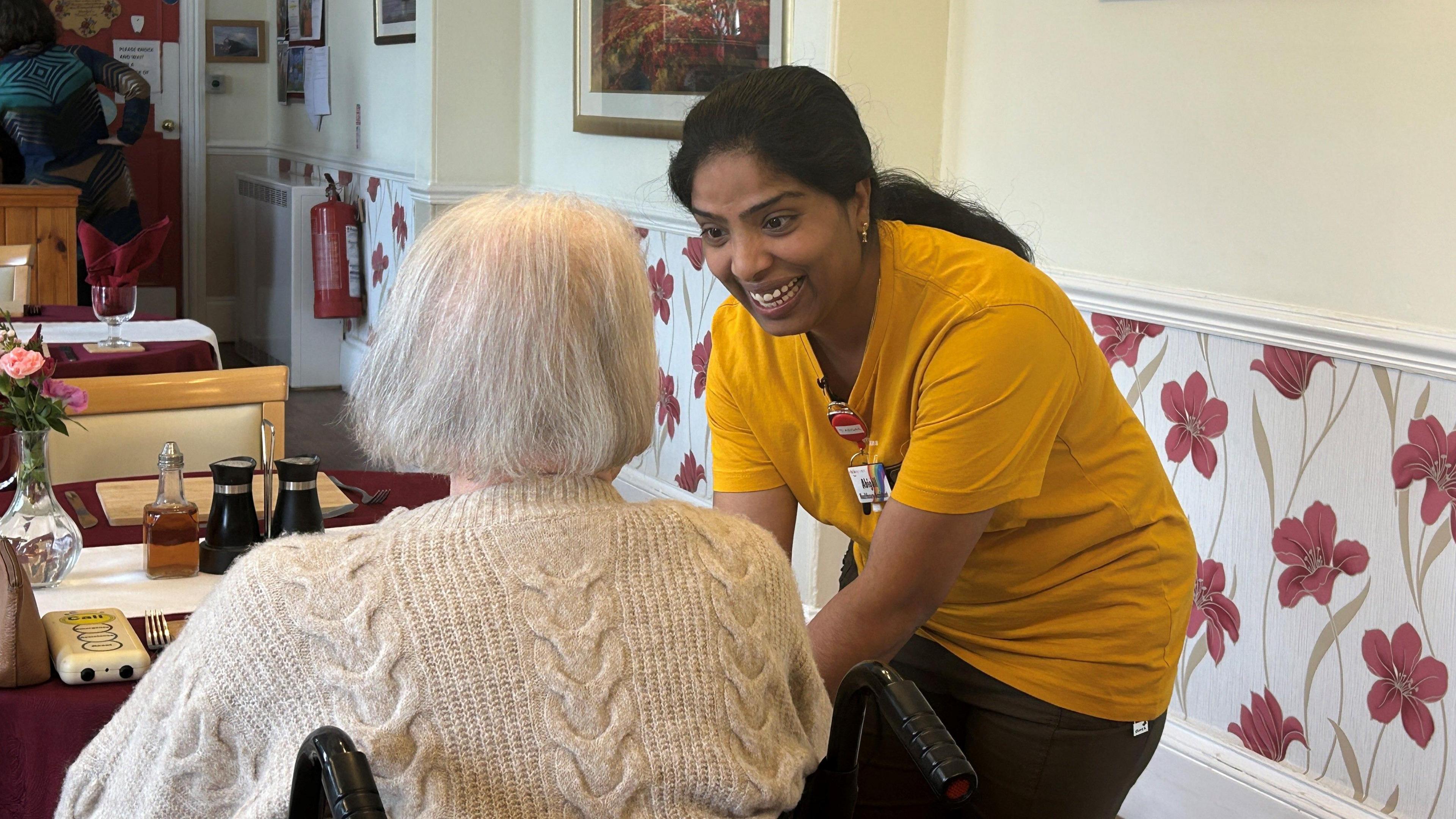 A woman dressed in a yellow T-shit leaning over an elderly lady sat in a wheelchair in a care home