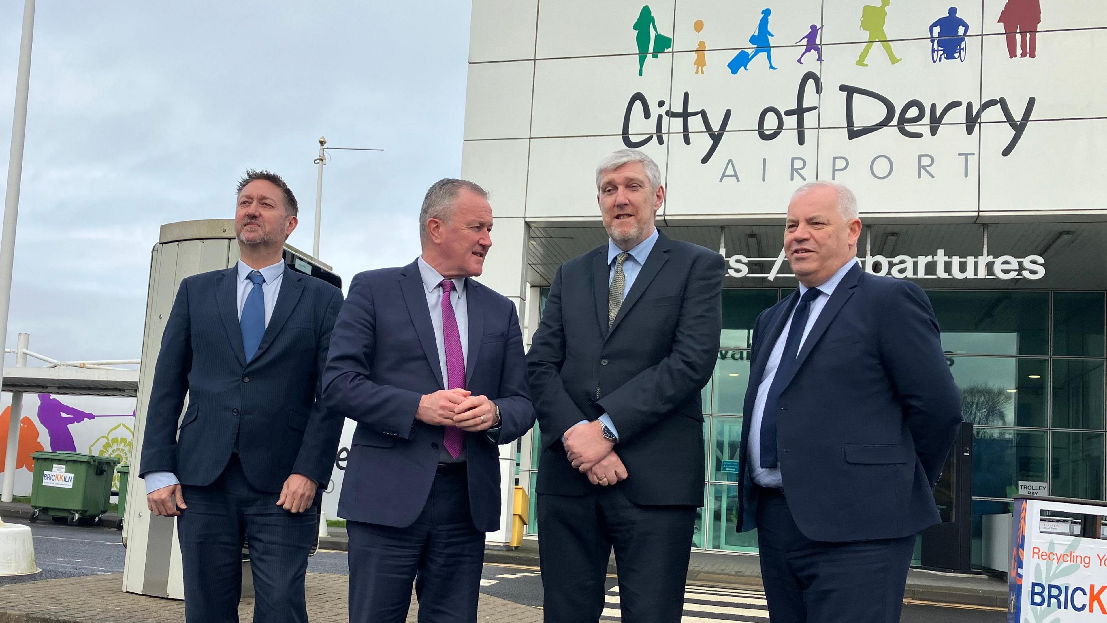 Four men standing outside the city of Derry airport. All men are dressed in navy suits and posing for a photograph.