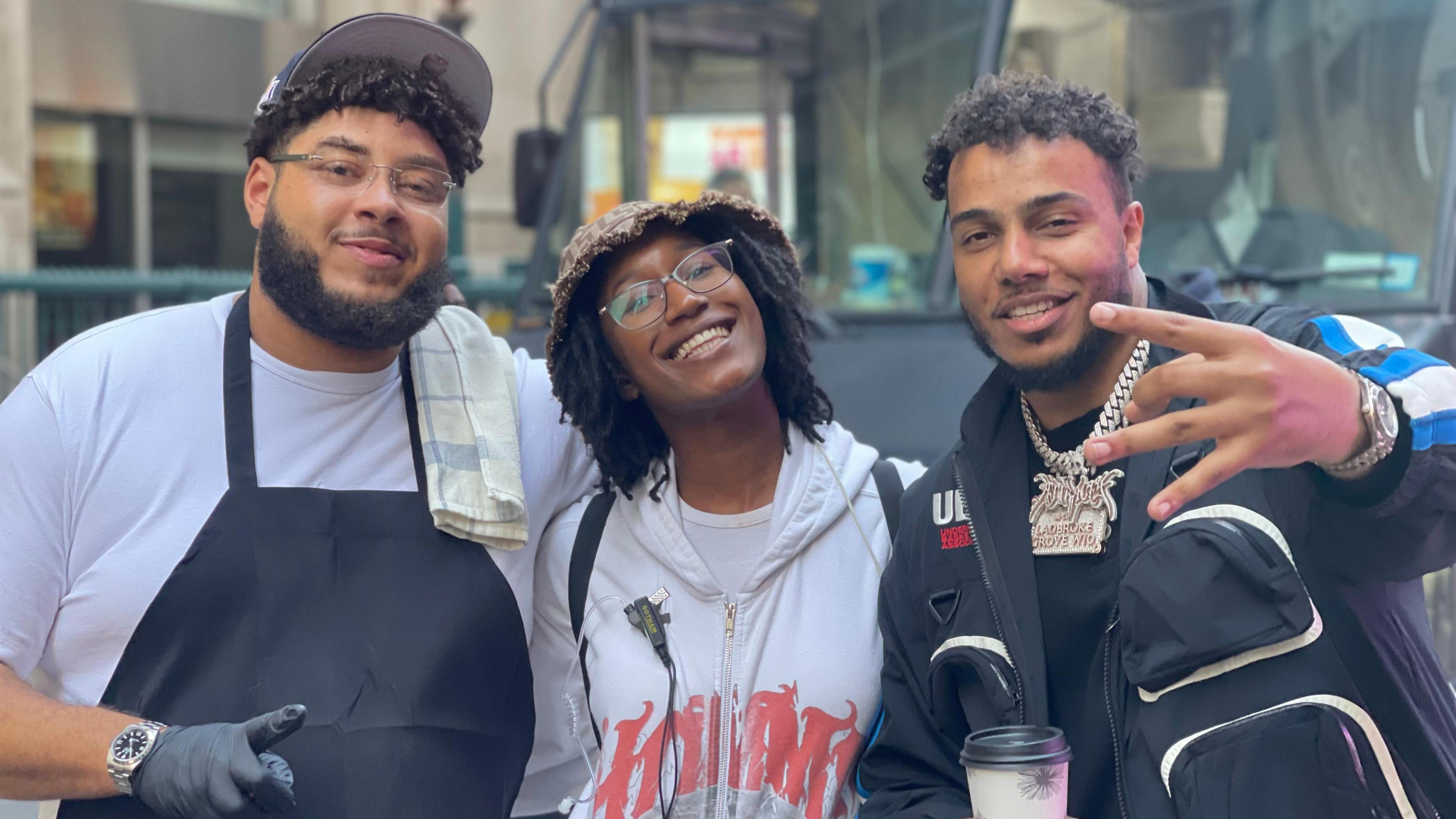 Big Zuu (left) is wearing a black apron over a white t-shirt, a grey cap, black gloves, and glasses, with a towel over his shoulder. Thaliqua Smith (centre) is smiling brightly, wearing glasses, a brown bucket hat, a white hoodie with red print, and a backpack. AJ Tracey (right) is in a black tracksuit with blue and white details, layered gold chains, and is holding a coffee cup while making a hand gesture. They're standing in front of a food truck on a city street.