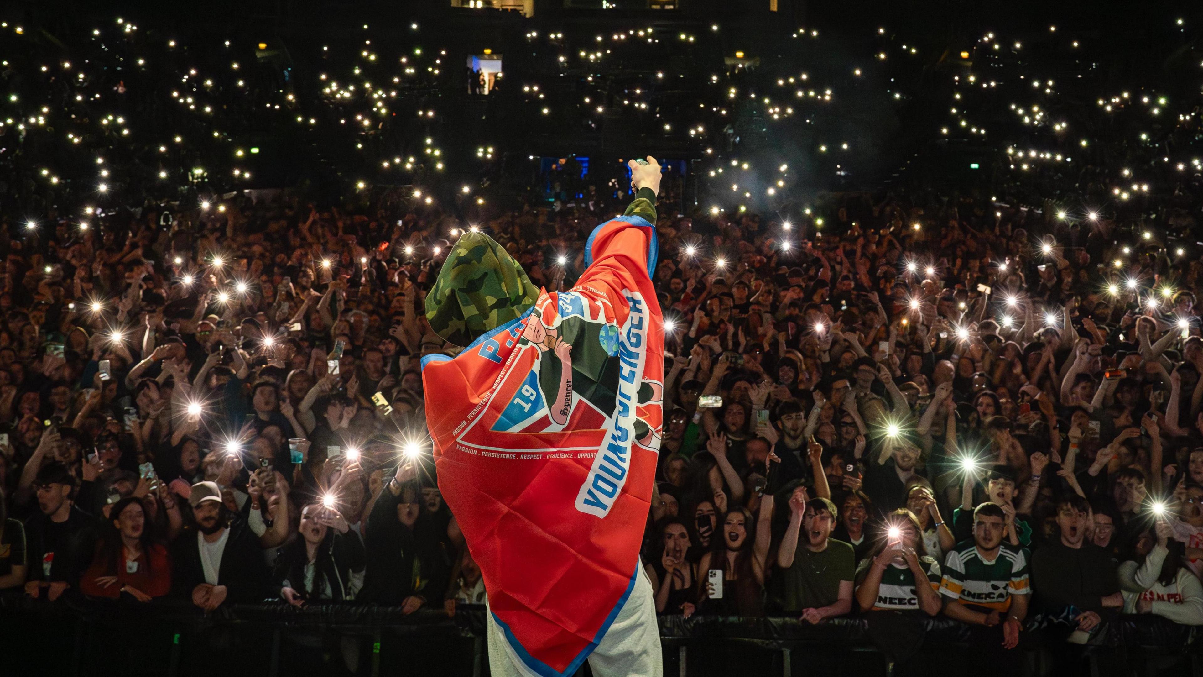 A man with a camo hoodie has his back to the camera whilst he faces a crowd of people as he performs on a stage. Some of the 100's of people in the crowd are filming him, lots of flashes can be seen as the crowd film him. The man wears a red flag across his back.