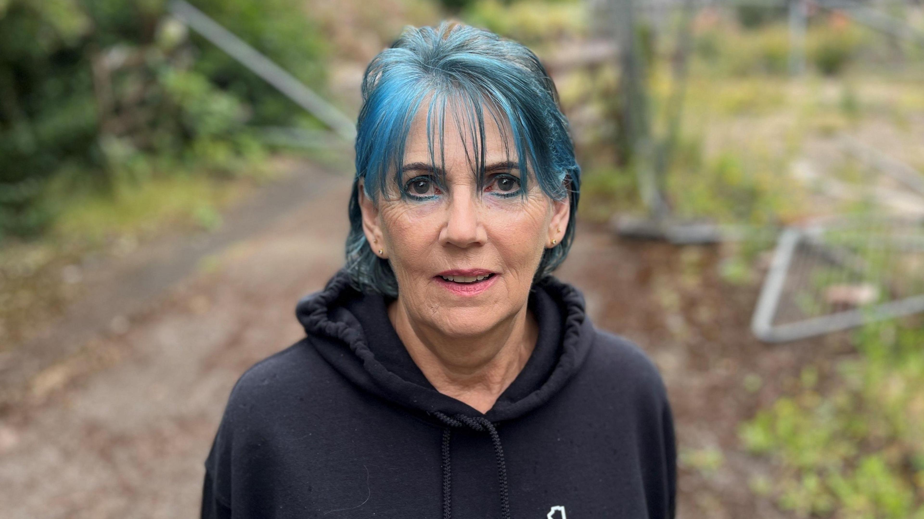 A woman with blue hair and a dark top is looking at the camera. In the background are fences and bushes that have been blurred.